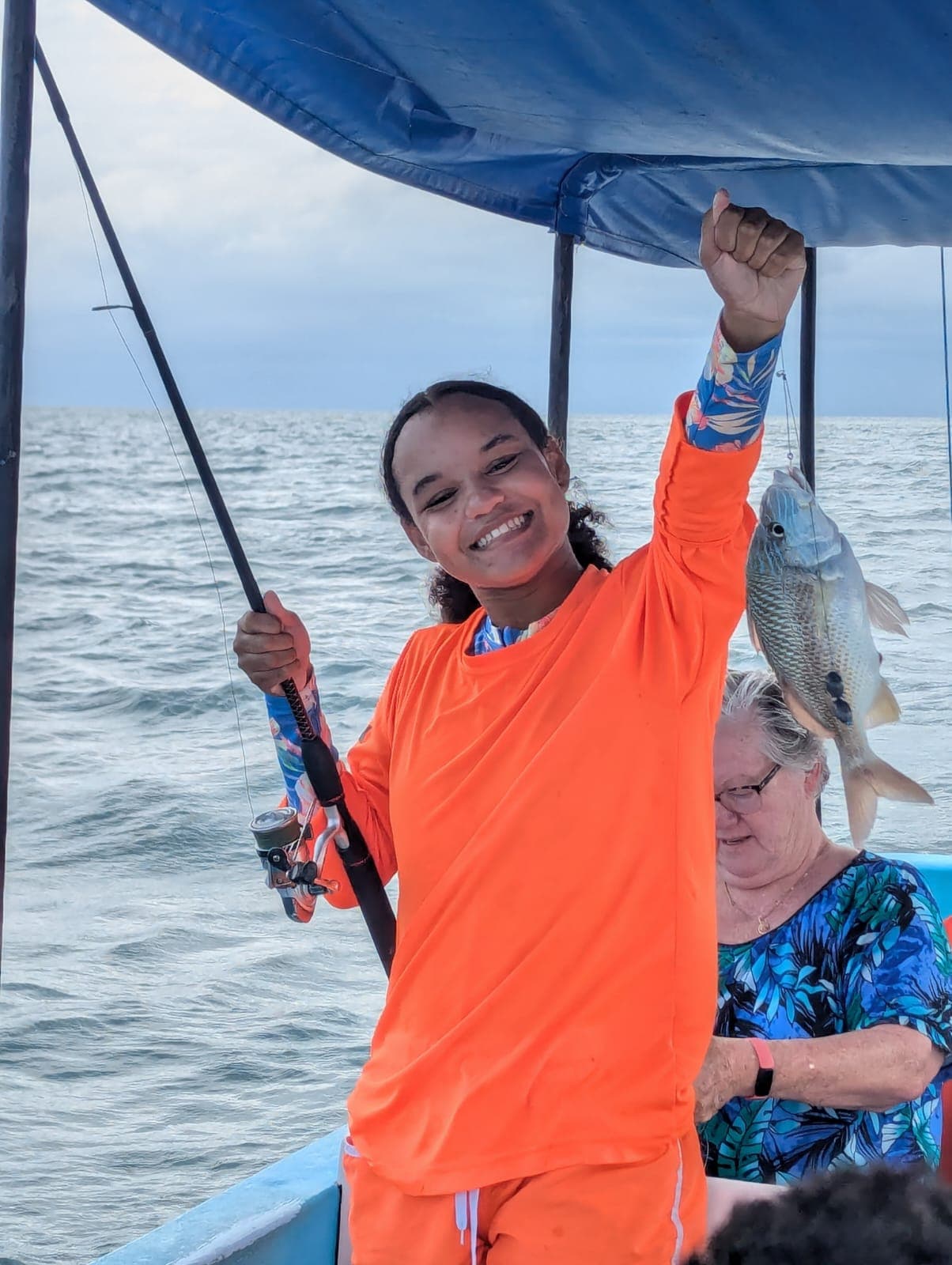 A smiling young woman in orange holds a fish on a fishing boat, with an older woman seated behind her.