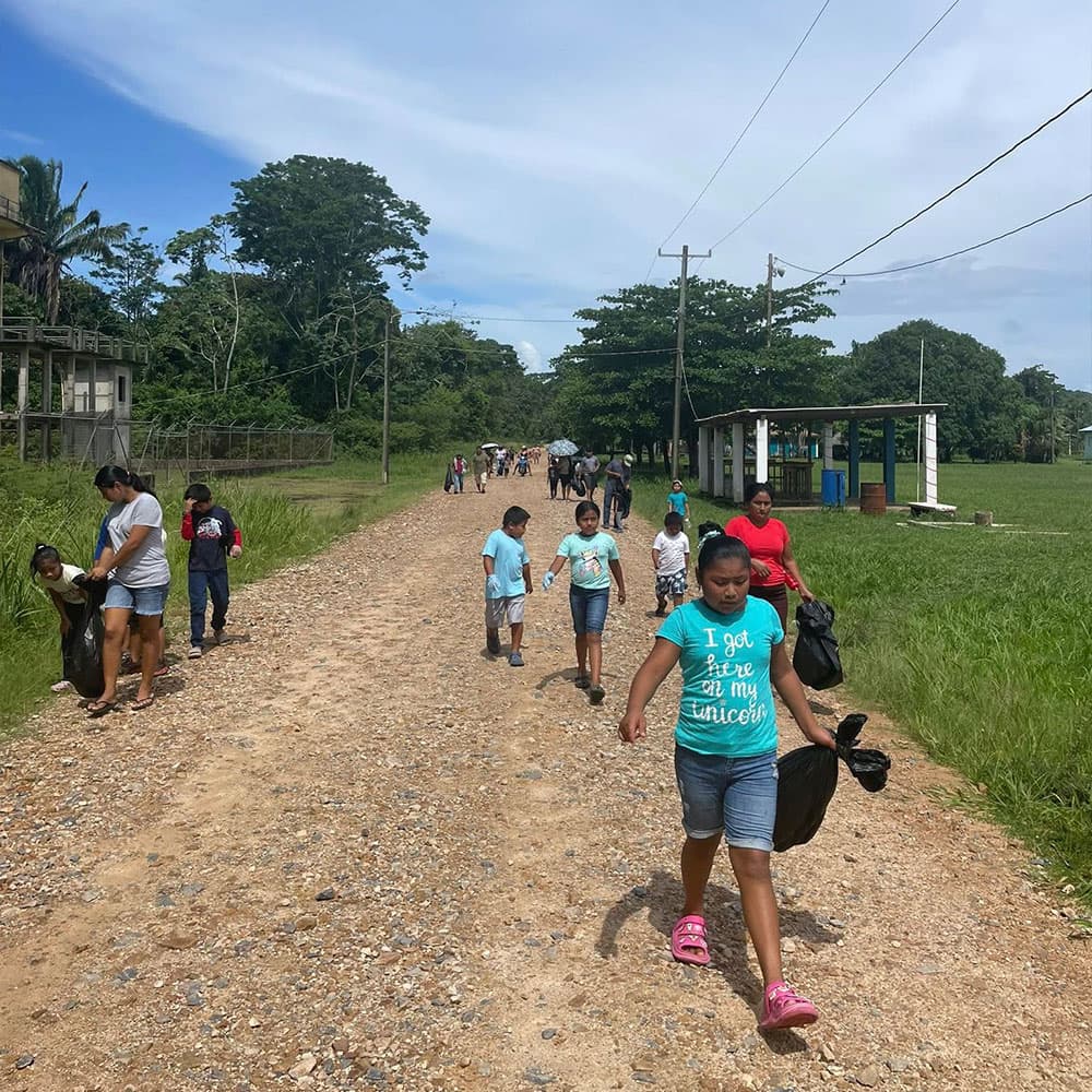 A group of children and adults walk down a gravel road lined with greenery and buildings under a blue sky.