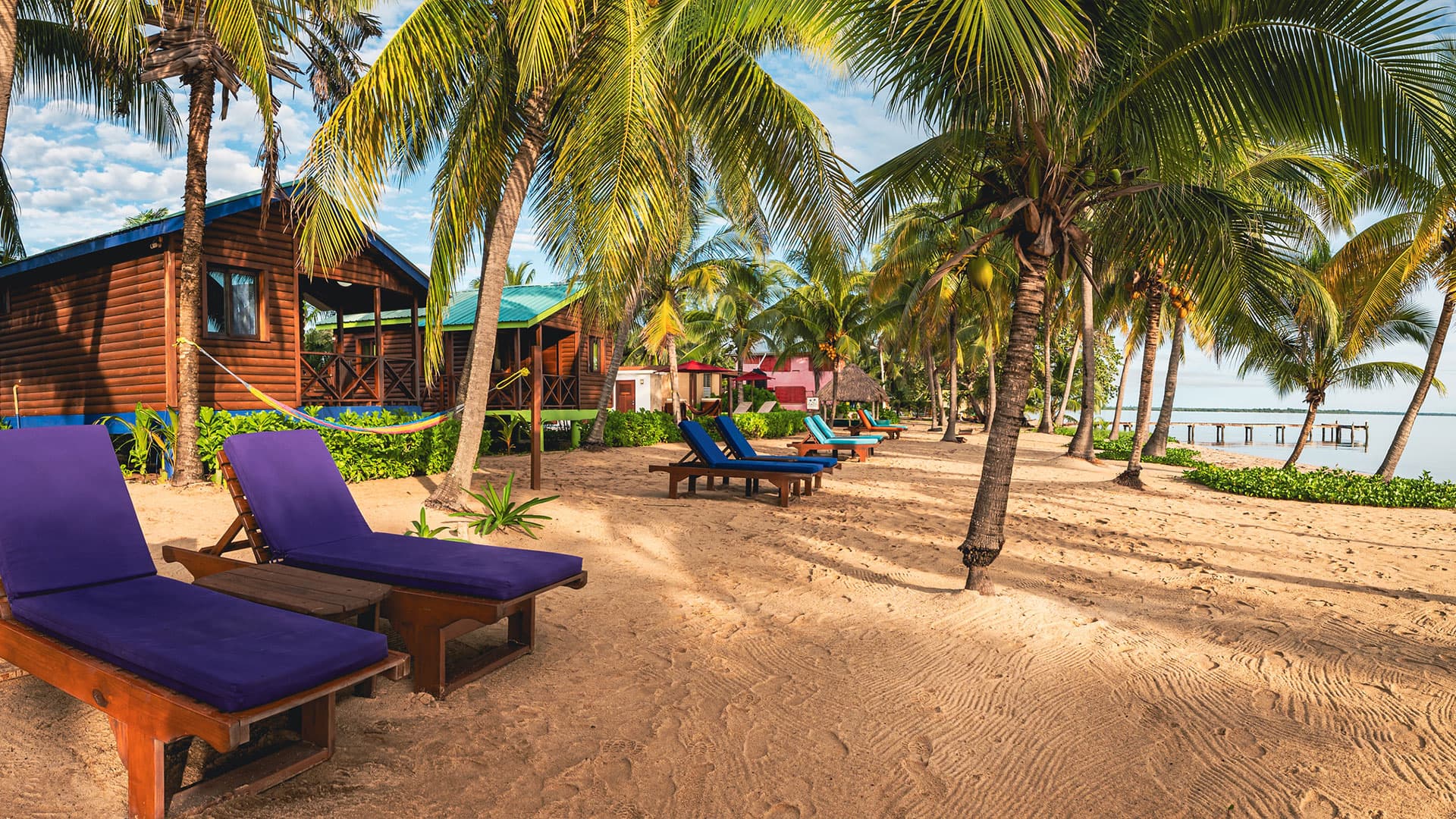 A sandy beach lined with colorful lounge chairs and palm trees, featuring wooden cottages in the background.