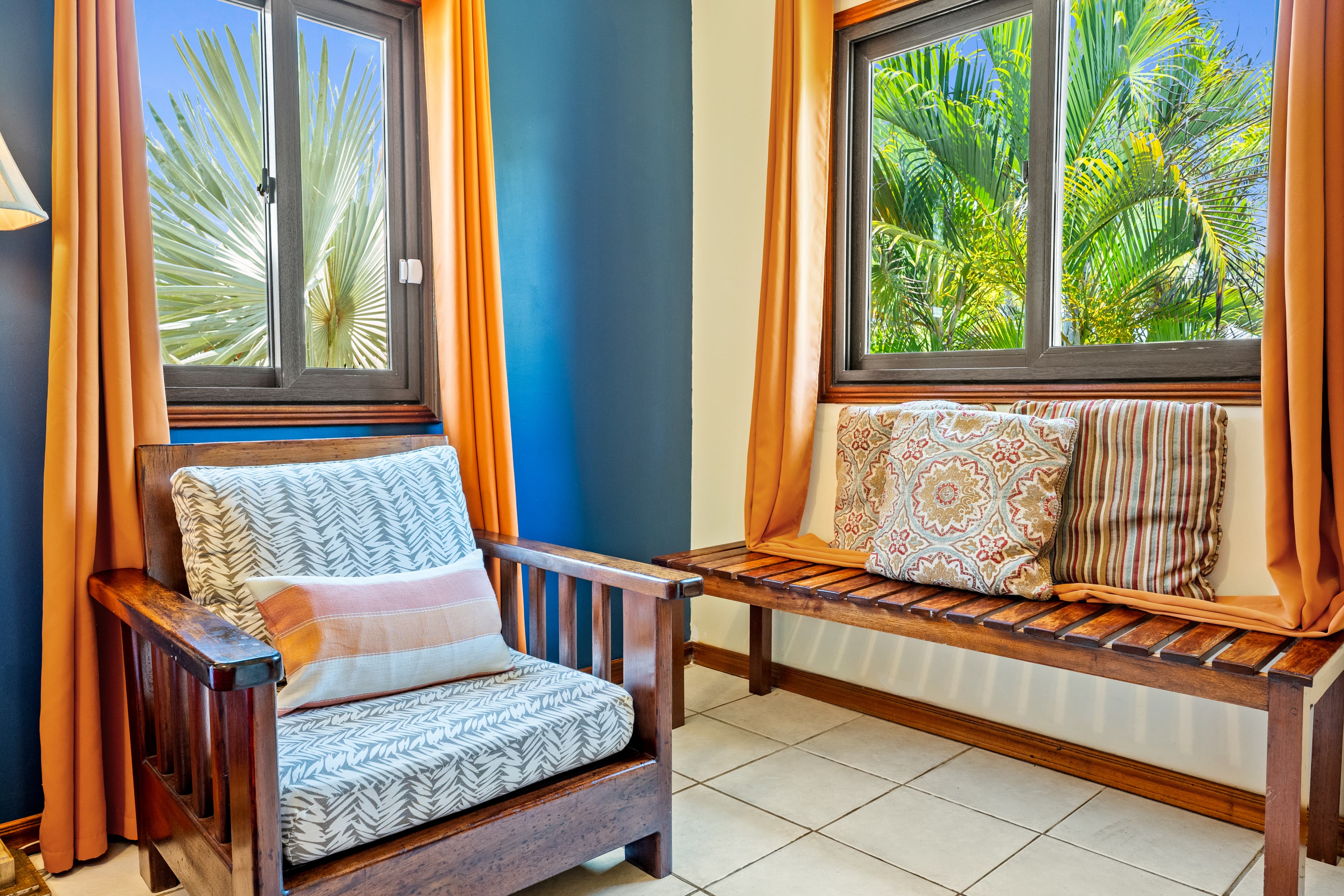 A cozy seating area in a vibrant bedroom featuring a dark wood armchair with patterned cushions and a matching wooden slatted bench. The space is framed by a deep blue accent wall and windows with orange curtains that look out onto lush tropical palm trees.