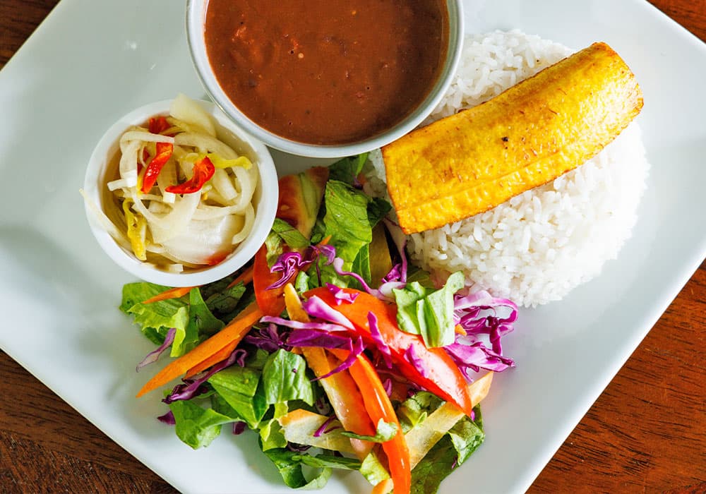A plate featuring rice, a fried plantain, salad, and a small bowl of beans with pickled vegetables.