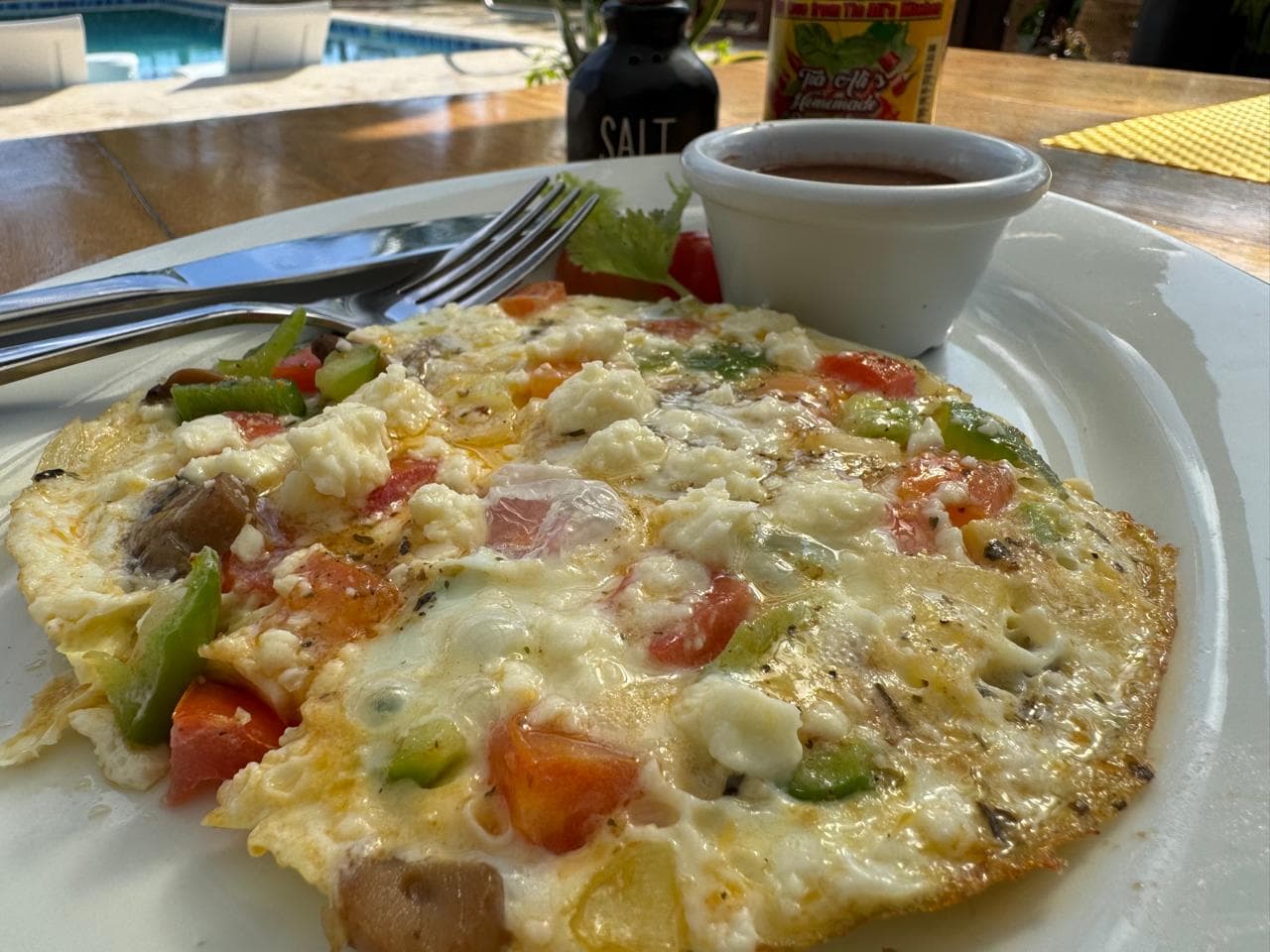 A colorful vegetable omelet served on a plate with a small cup of sauce and a bottle of salt in the background.