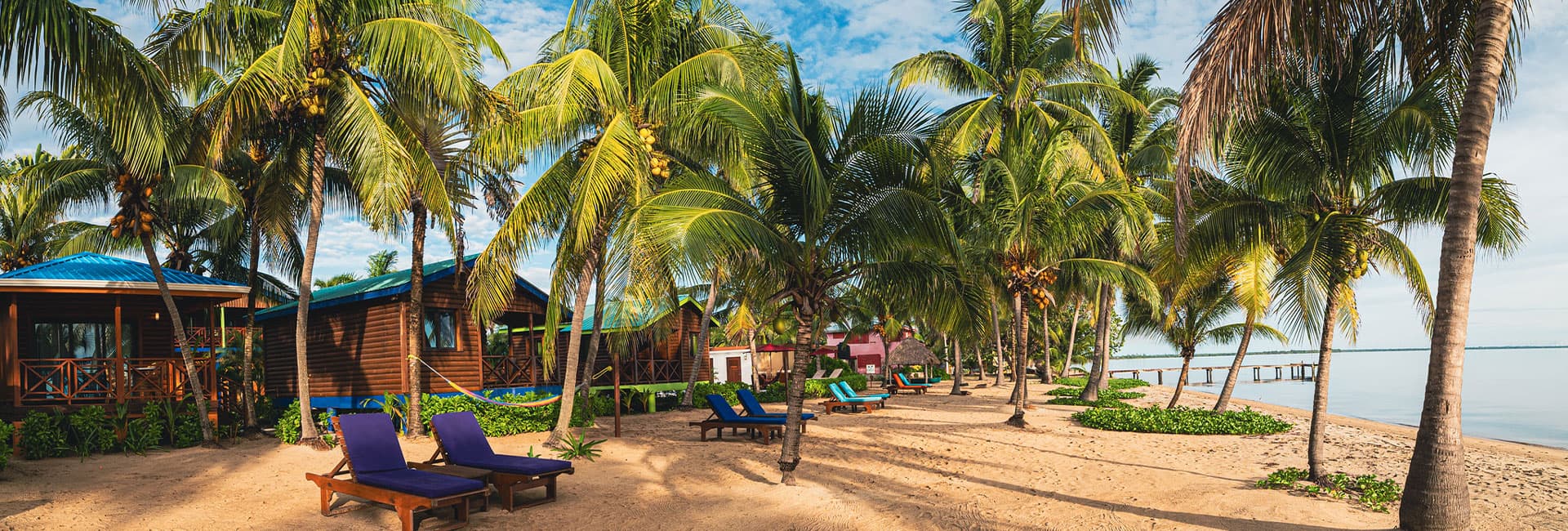 A tropical beach scene with palm trees, wooden cottages, and lounge chairs by the shore.