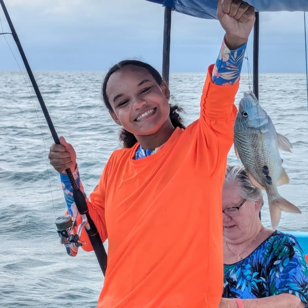 A person in an orange shirt joyfully holds up a fish while another person sits behind them on a boat.
