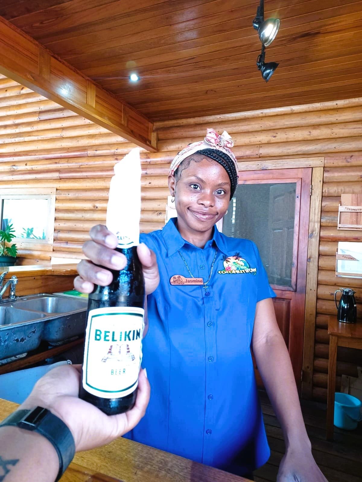 A woman in a blue shirt holds up a bottle of Belikin beer with a smile.