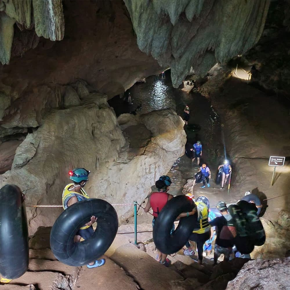 A group of adventurers descends into a cave, carrying inner tubes near a stream.