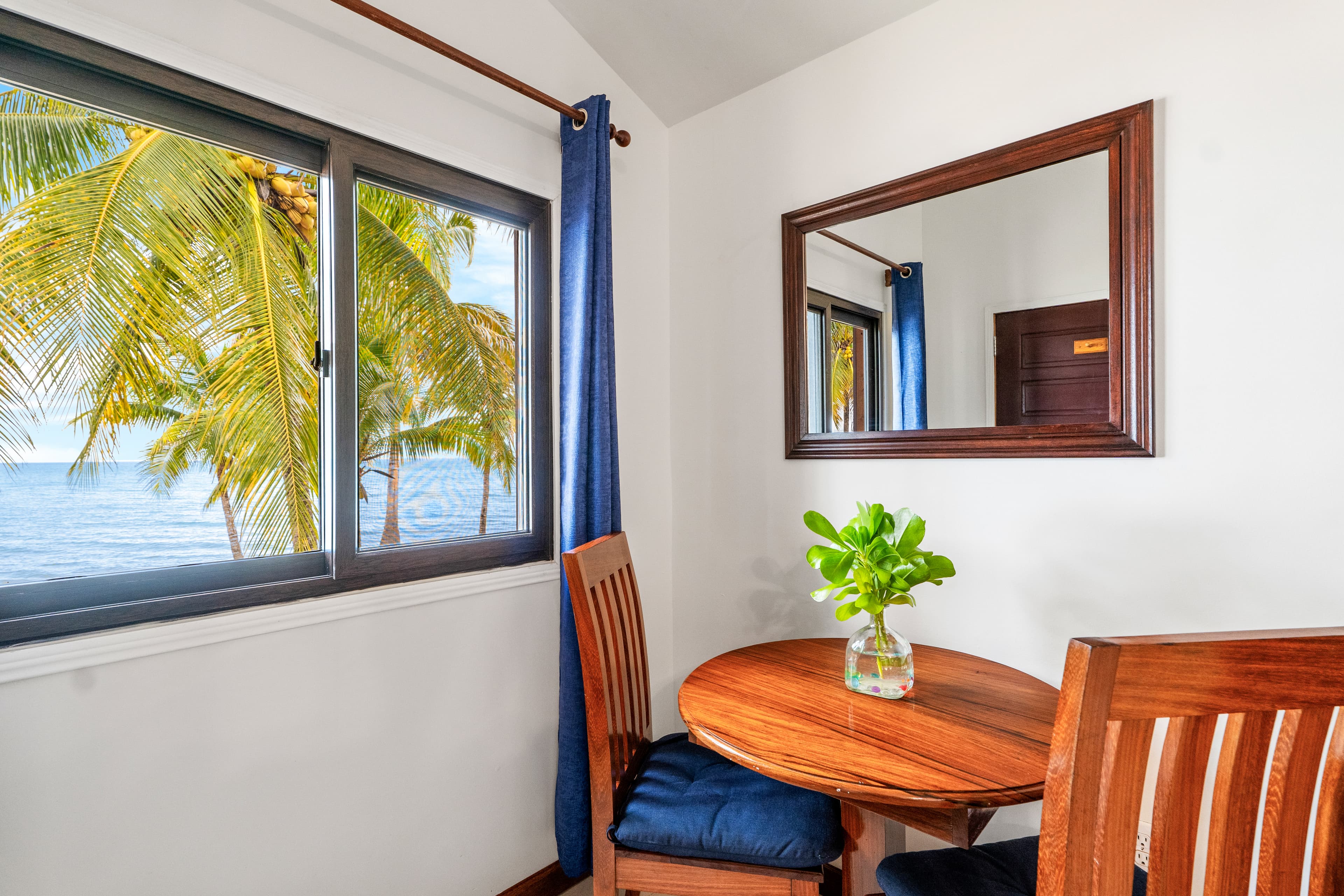 A simple wooden dining table for two is set against a white wall with a dark wood-framed mirror. The table is decorated with a small glass vase of green foliage and is flanked by two chairs with blue cushions, while a nearby window with blue curtains offers a view of a palm-fringed tropical beach.