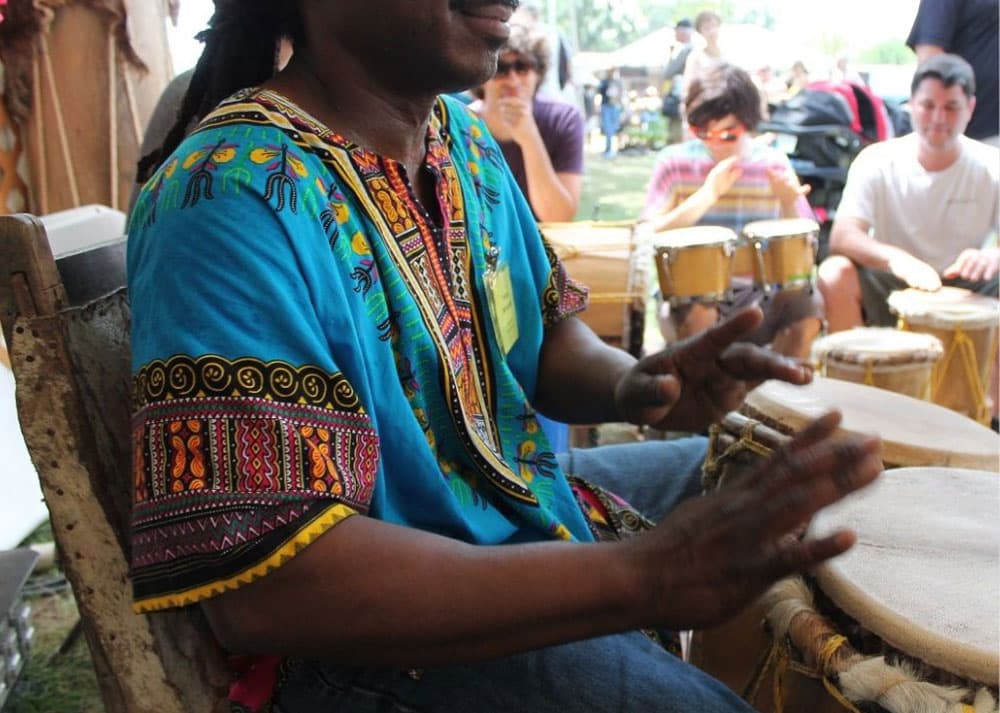 A man in a colorful shirt plays a drum while others enjoy the music in the background.