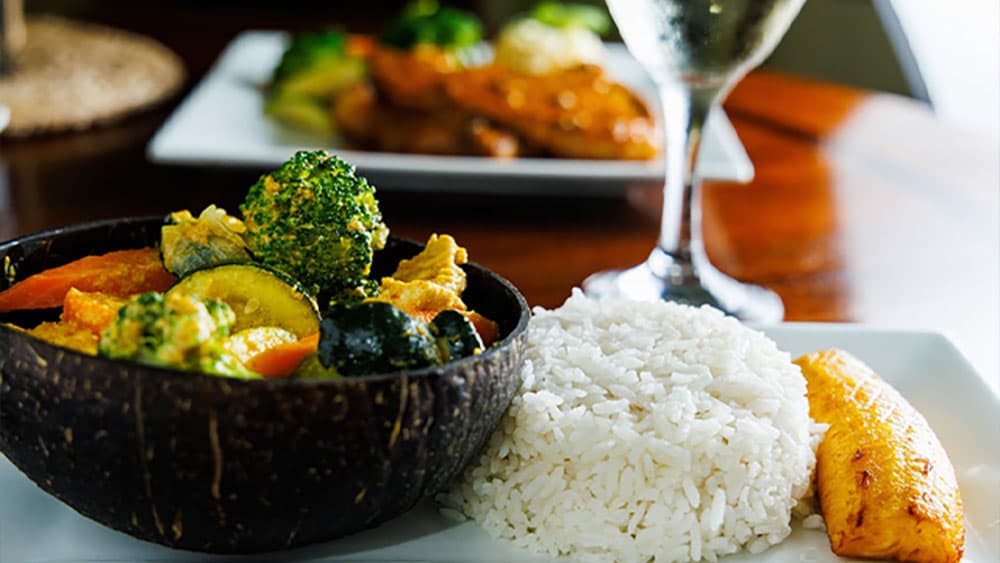 A coconut bowl filled with mixed vegetables alongside a serving of rice and a side of fried plantains.