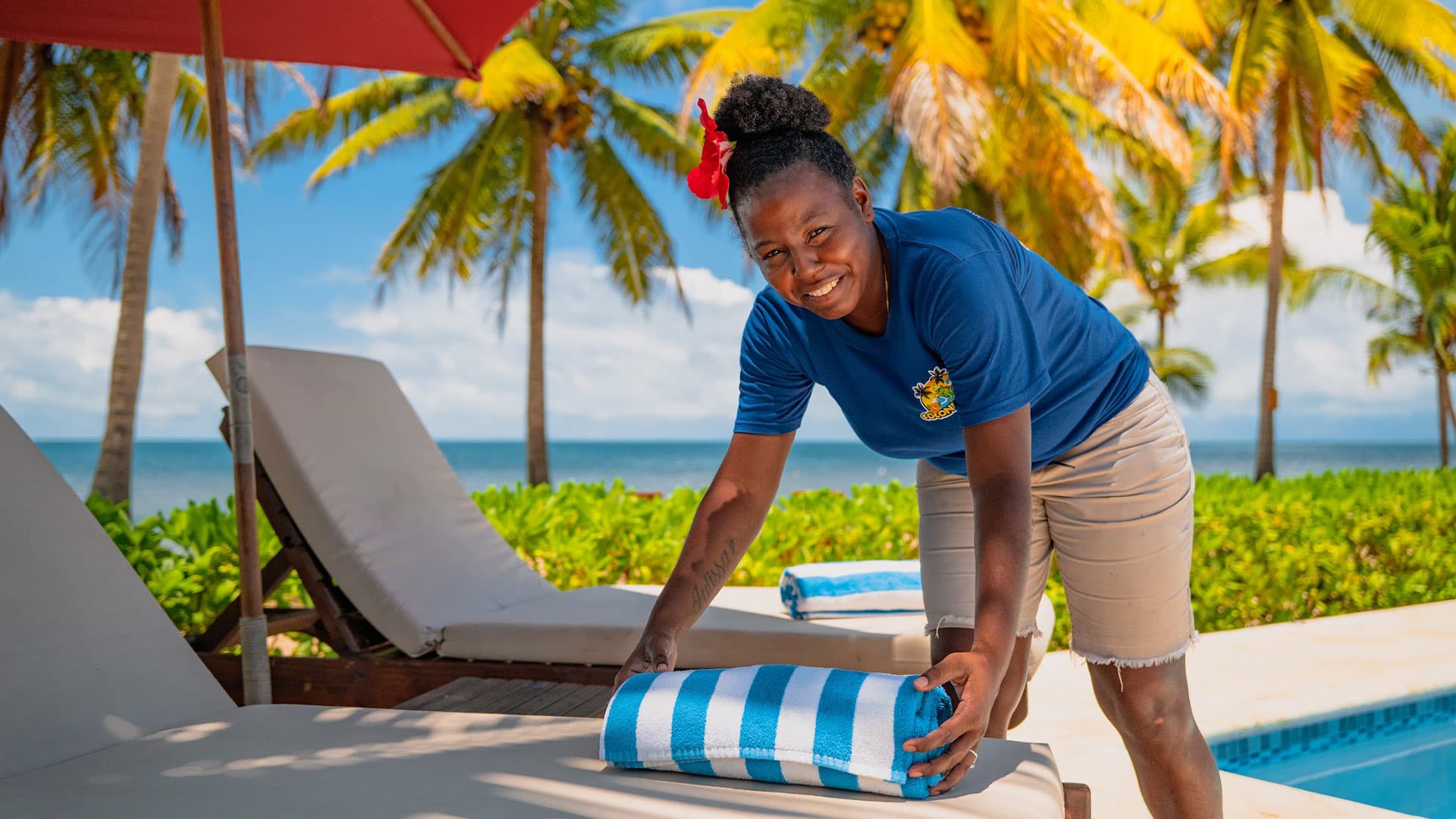 A smiling woman in a blue shirt arranges a striped towel on a lounge chair by a pool surrounded by palm trees.