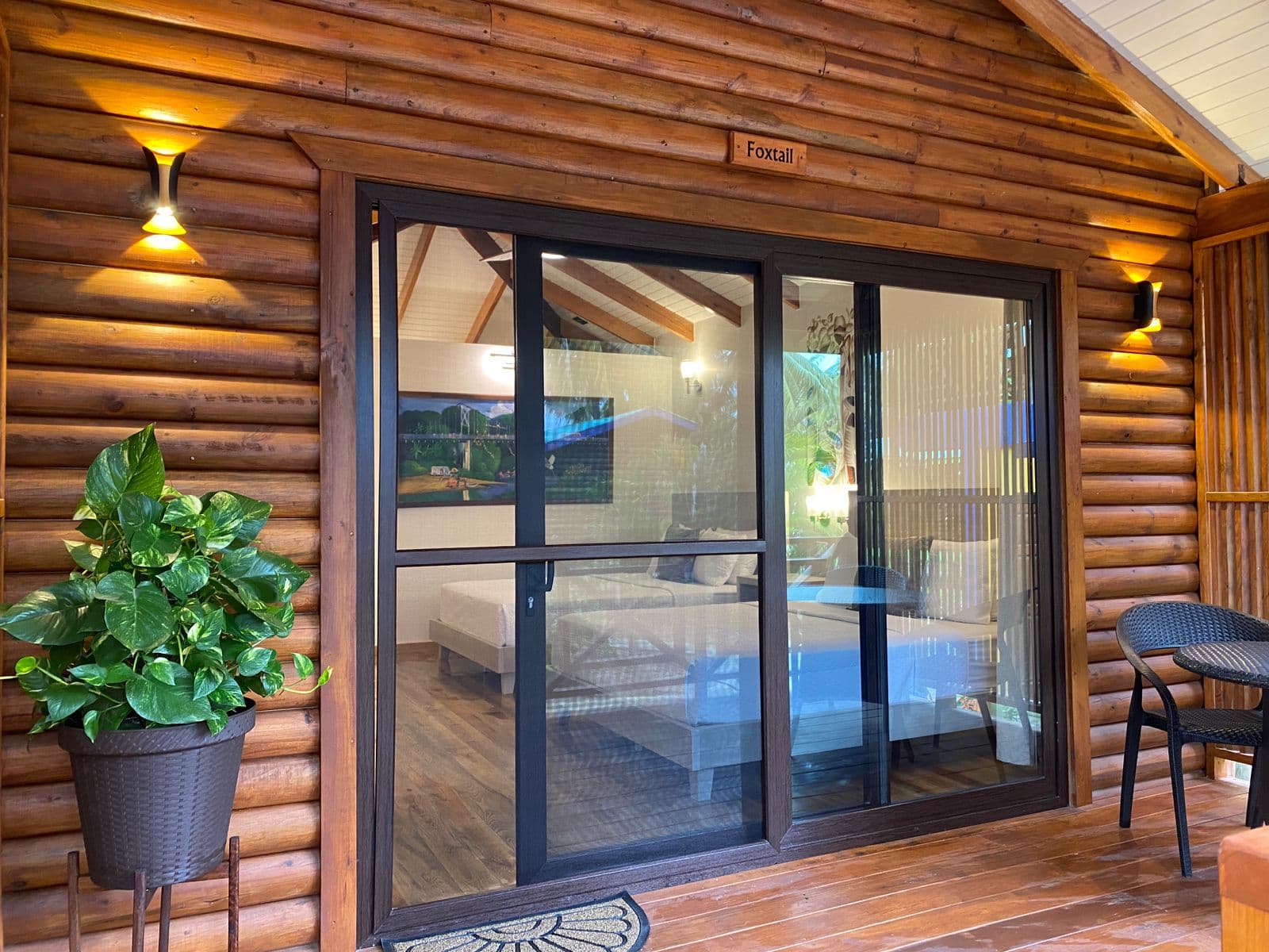 A cabin exterior features large glass sliding doors that provide a clear view into a bedroom with two beds. To the left of the doors, a large green potted plant sits on the wooden porch, illuminated by a warm, hourglass-shaped wall light.