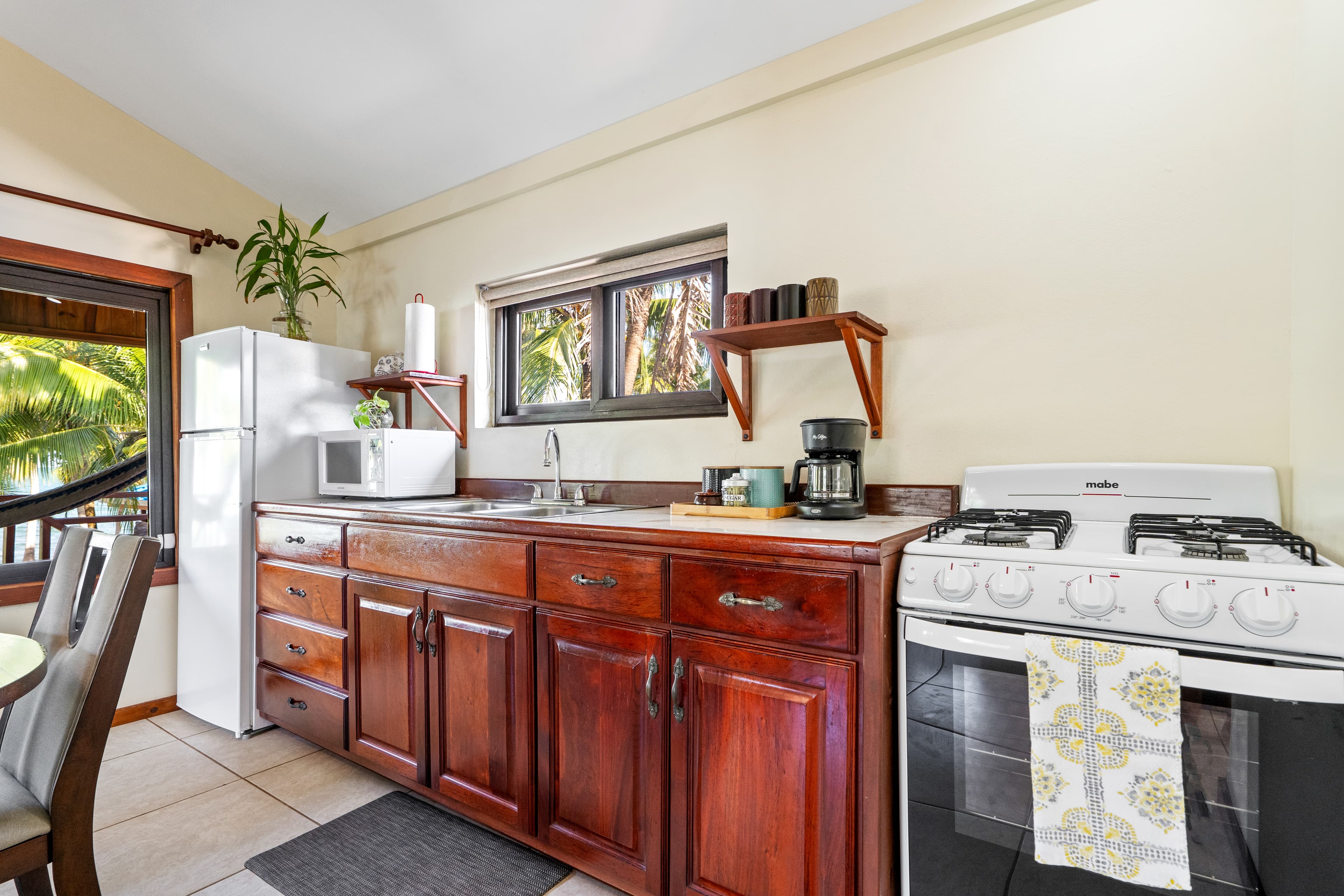 A kitchen area featuring dark wood cabinetry and a white countertop with an integrated sink. The space is equipped with a white refrigerator, a microwave, a coffee maker on a wooden shelf, and a white gas stove with a patterned towel hanging from the oven handle. A window above the sink looks out onto tropical greenery.