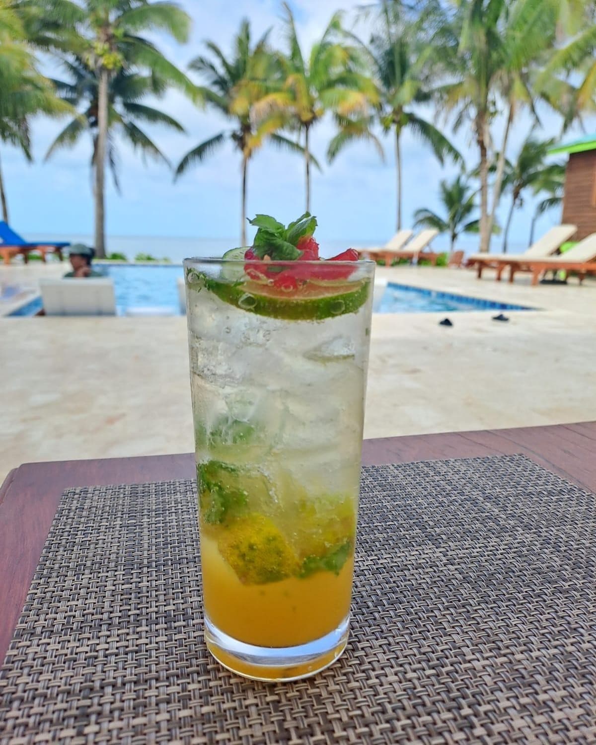 A refreshing cocktail with lime and mint sits on a table, overlooking a tranquil pool surrounded by palm trees.