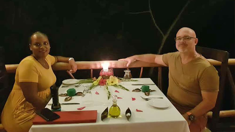 A couple sits at a beautifully set table for two, illuminated by a candle, in a romantic outdoor setting.