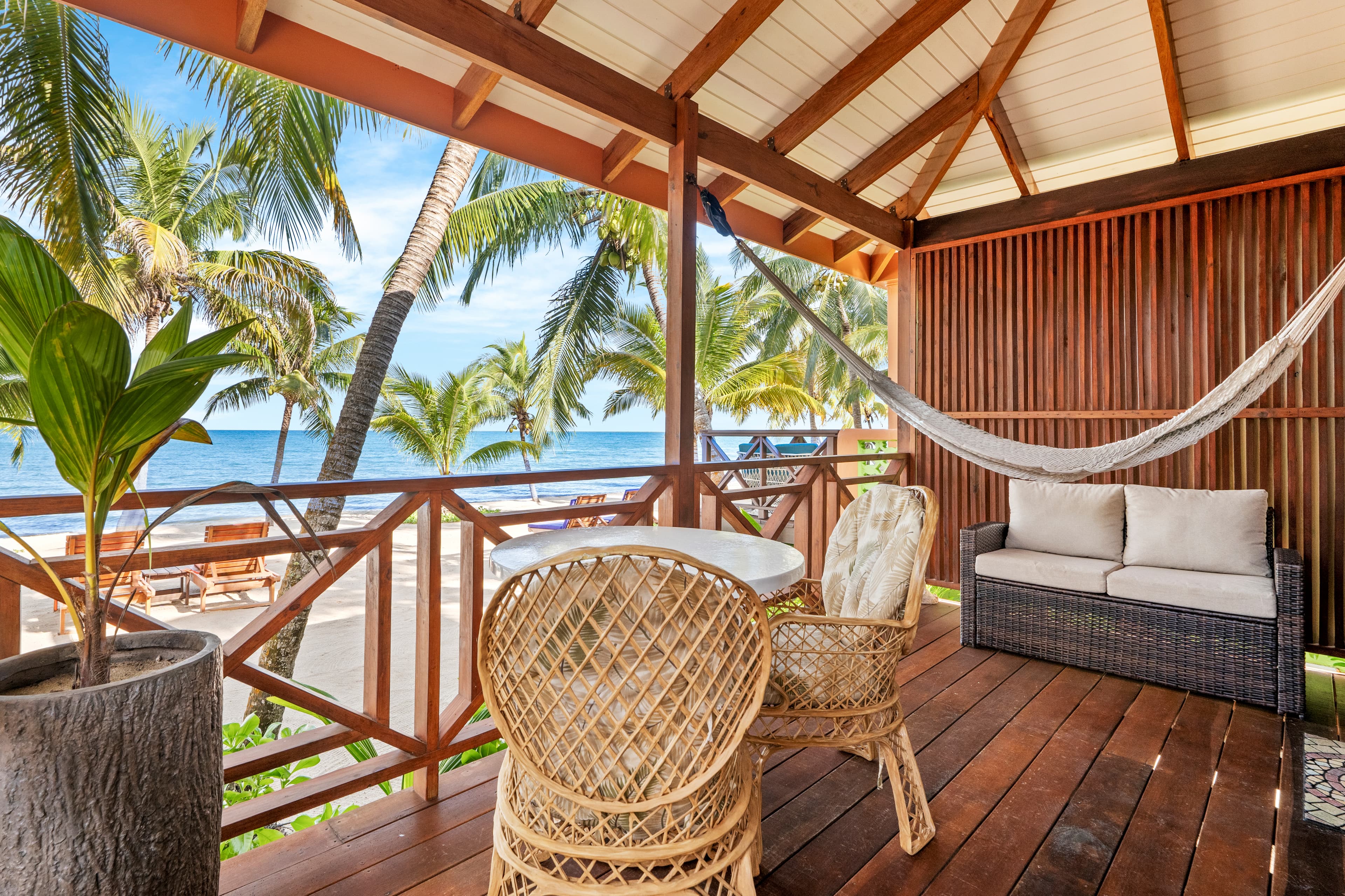 A spacious wooden balcony with a vaulted ceiling and a polished wood floor features a seating area with a small white table, two wicker chairs, and a dark wicker loveseat with white cushions. A white woven hammock is strung across the space, and a large potted plant sits in the foreground, all overlooking a tropical beach with palm trees and the sea.