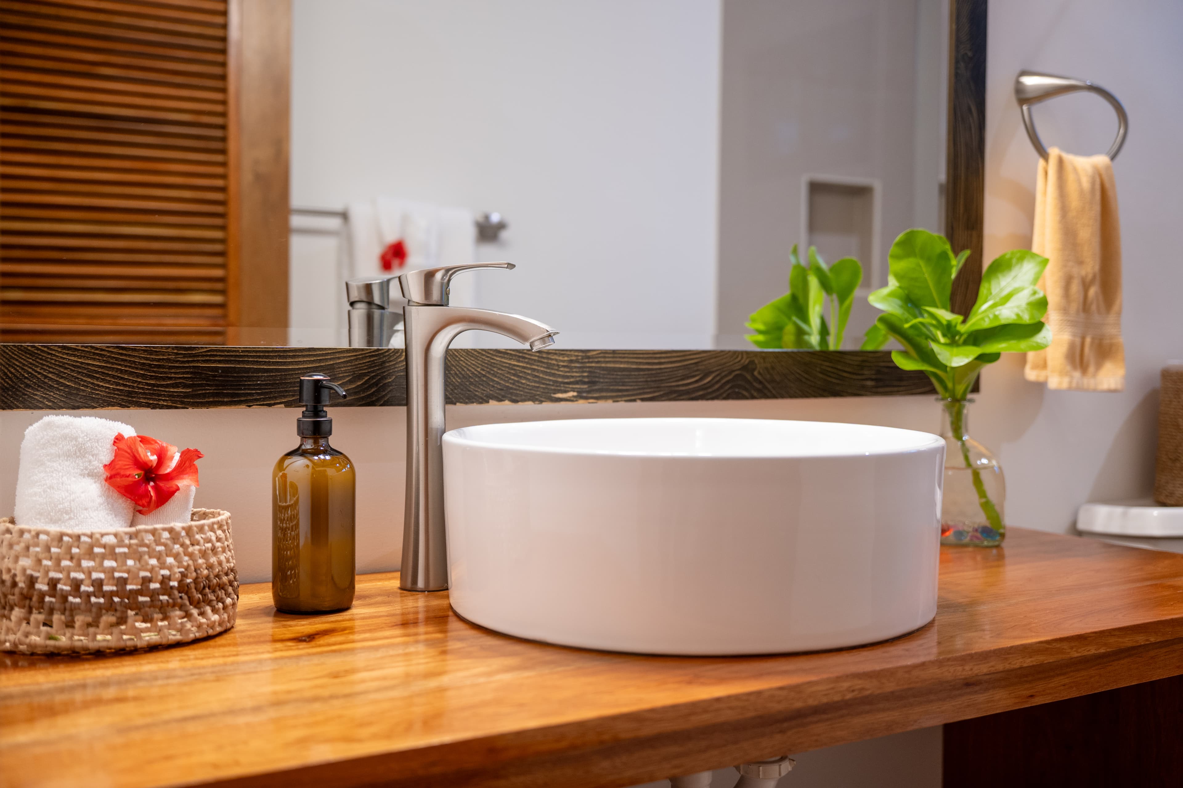 A modern bathroom featuring a white vessel sink on a dark wood-grain vanity with a matching wood-framed mirror. The space includes a walk-in shower with a glass partition and a decorative mosaic-tiled accent wall in shades of blue and tan. On a black shelf above the toilet, there is a white vase with dried pampas grass and a small potted plant.