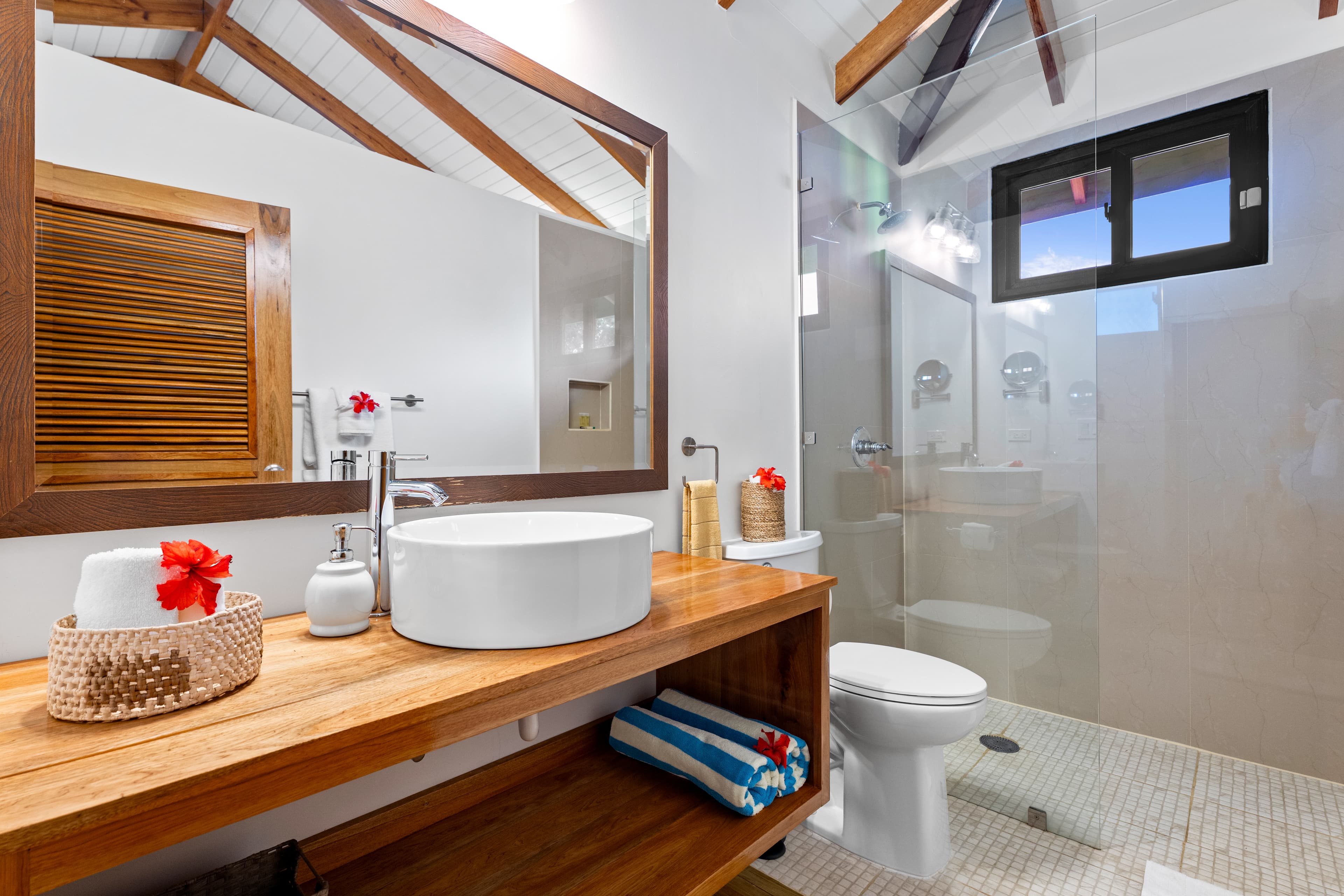 A modern bathroom with a white vessel sink on a wooden vanity featuring a large framed mirror. The room has a walk-in shower with a glass door and white pebbled flooring, neutral walls, and a vaulted ceiling with exposed wood beams.
