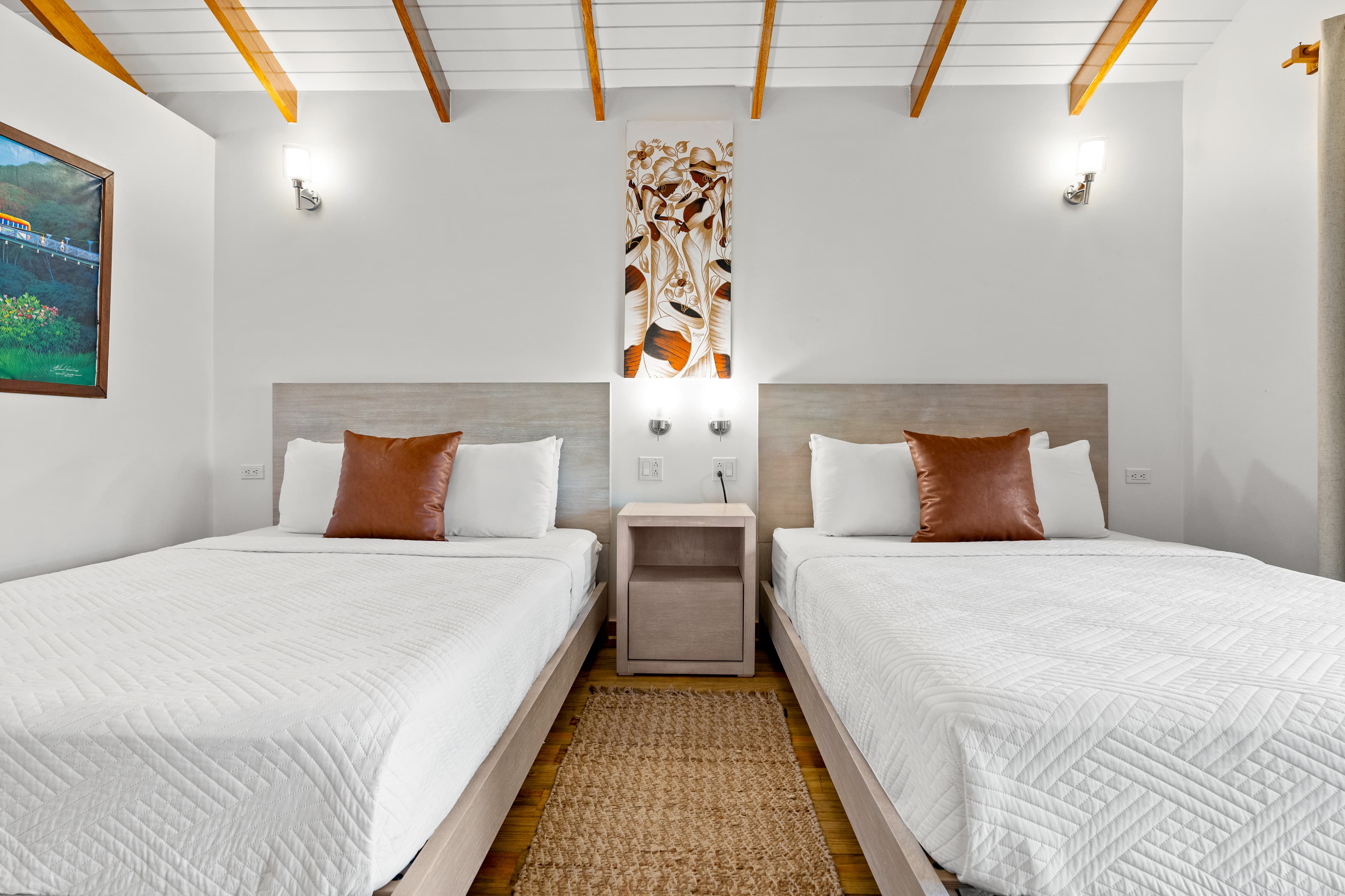 A bedroom with light-colored wood floors and a vaulted wood-beam ceiling features two queen beds with white linens and single brown accent pillows. A narrow woven rug lies between the beds, and a large piece of floral art hangs on the white wall above a shared nightstand.