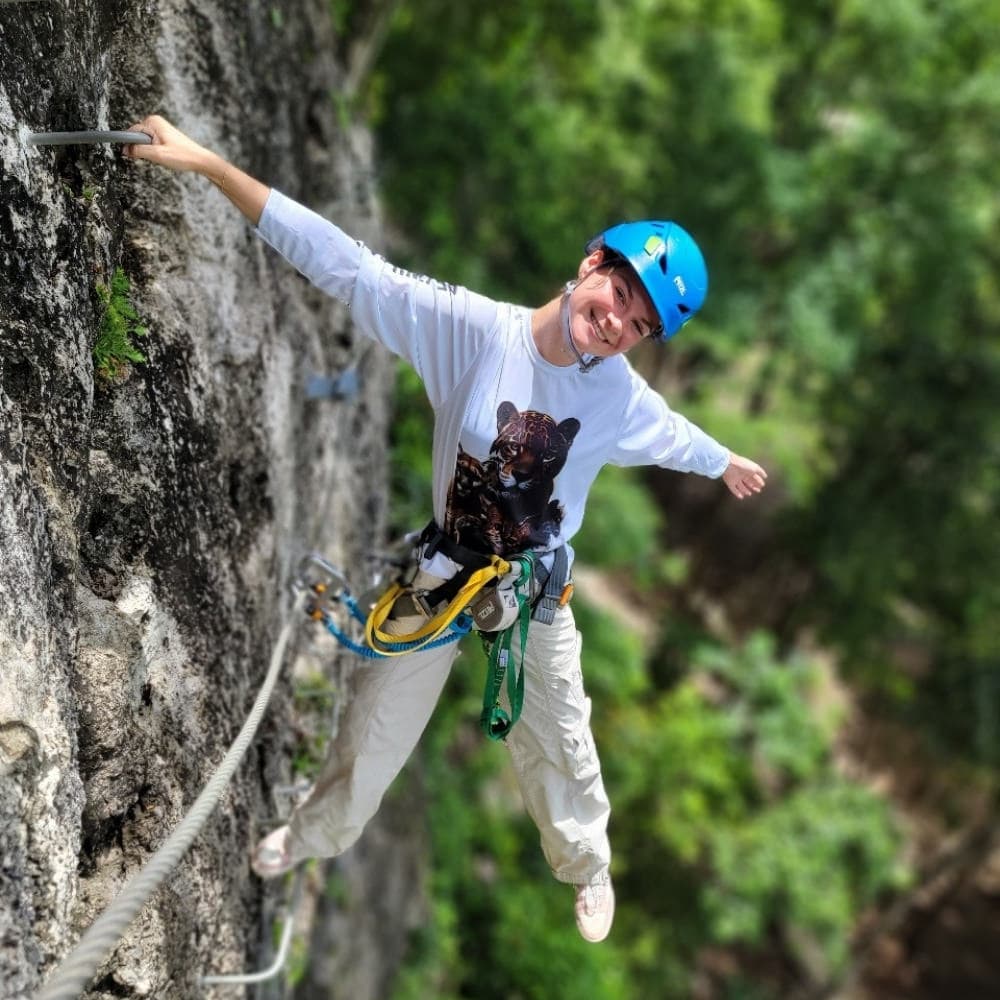 A smiling climber in a blue helmet poses on a rock face, harnessed with climbing gear against a lush green background.