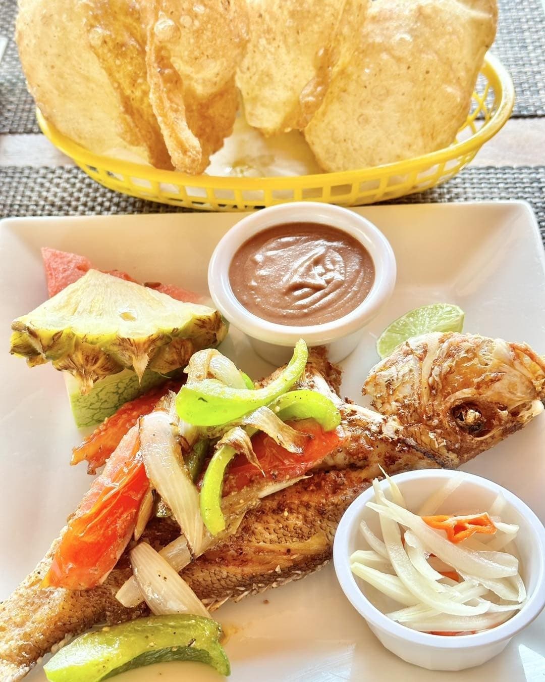 A plate of seasoned fried fish garnished with vegetables, accompanied by tropical fruit, dipping sauce, and crispy chips.