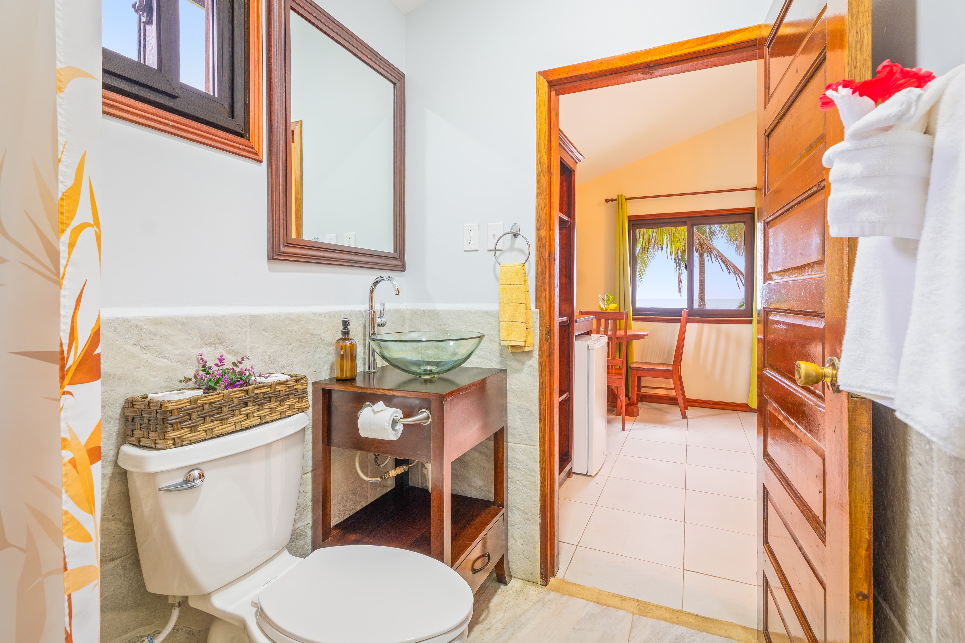 A compact bathroom featuring a white toilet on the left and a dark wood vanity with a glass vessel sink. The room has light-colored marble-patterned wall tiles and a view through an open wooden door into a bedroom area with a yellow bed runner and lime green curtains.
