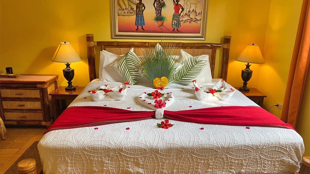 A beautifully arranged hotel room with a decorated bed featuring flowers, palm leaves, and a vibrant red runner against a cheerful yellow wall.