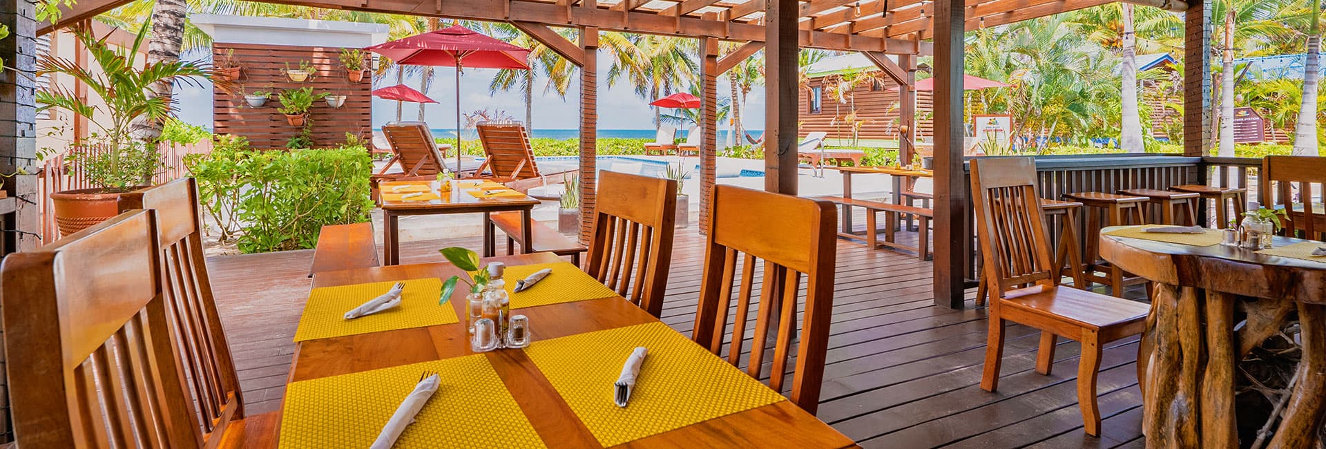A tropical dining area with wooden tables, yellow placemats, and views of palm trees and a pool.