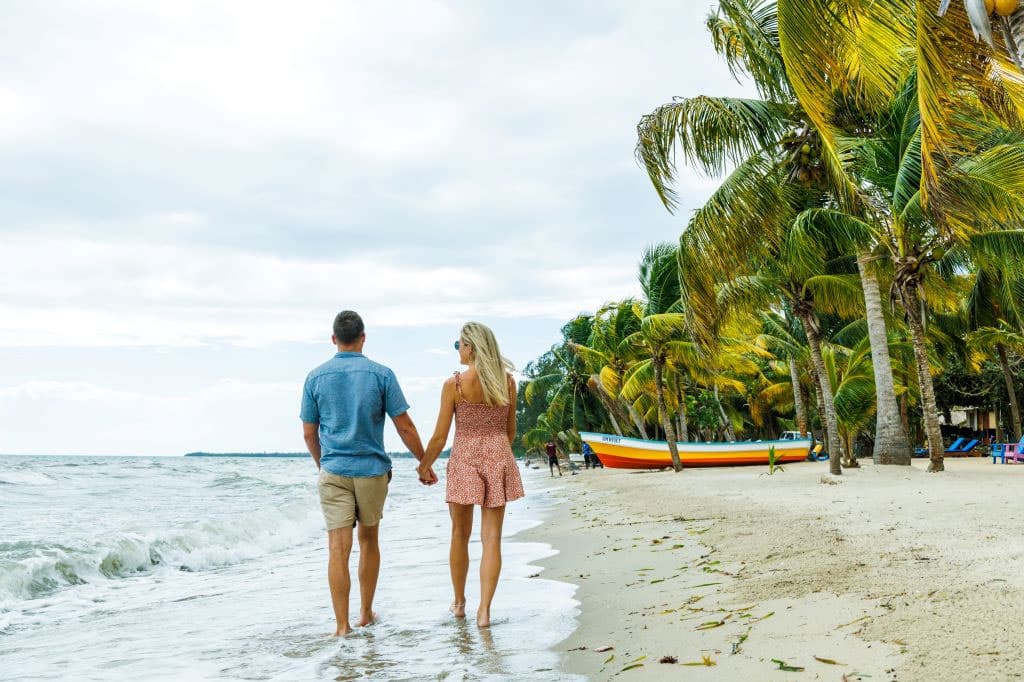 A couple holds hands while walking along a sandy beach lined with palm trees.