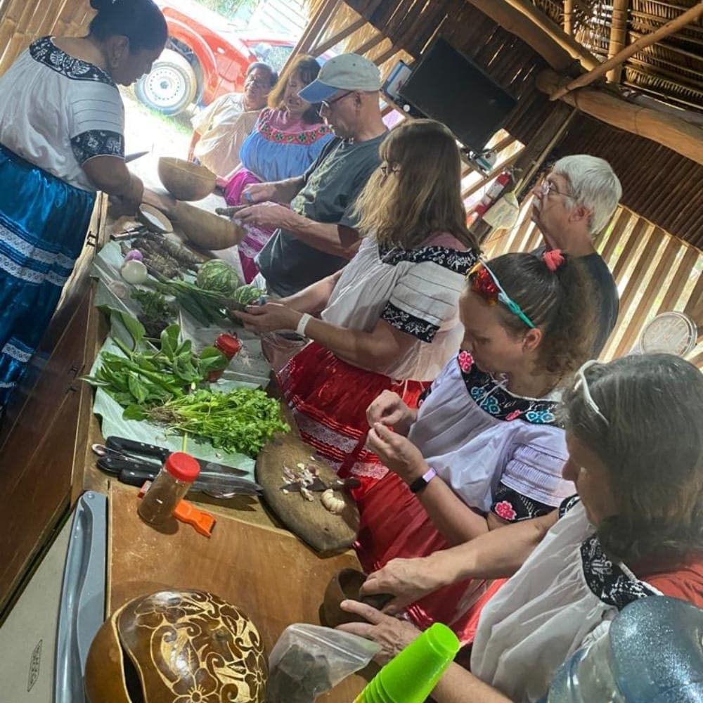 A group of people in traditional clothing prepare food at a wooden table filled with fresh ingredients.