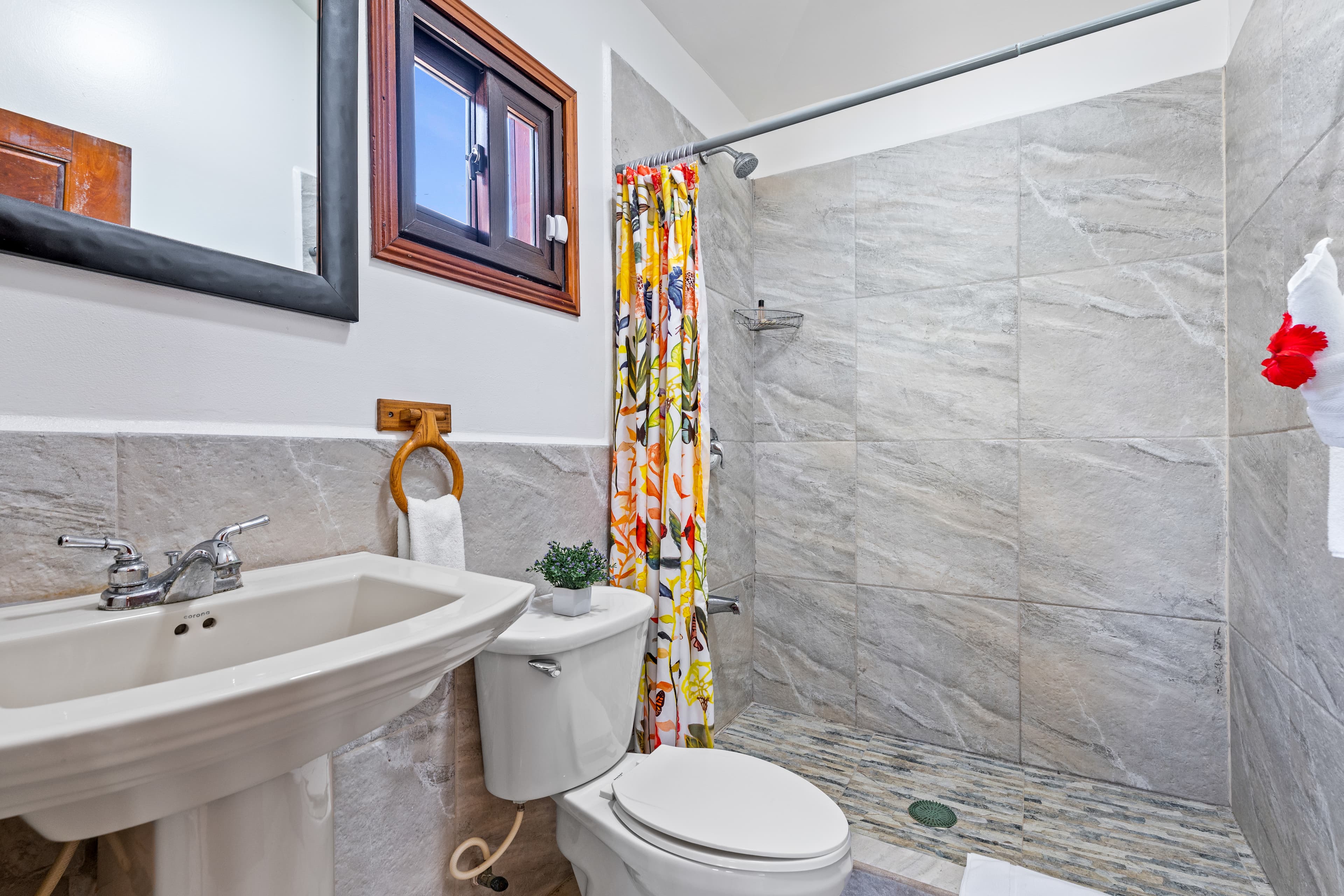 A bathroom with marble-patterned wall tiles and a walk-in shower featuring a vibrant, multi-colored floral curtain. The room includes a white pedestal sink, a white toilet, and a dark wood-framed mirror on the left, with a small window positioned above the toilet.