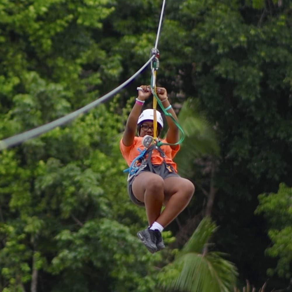 A person in an orange shirt zip-lines through a lush green forest.