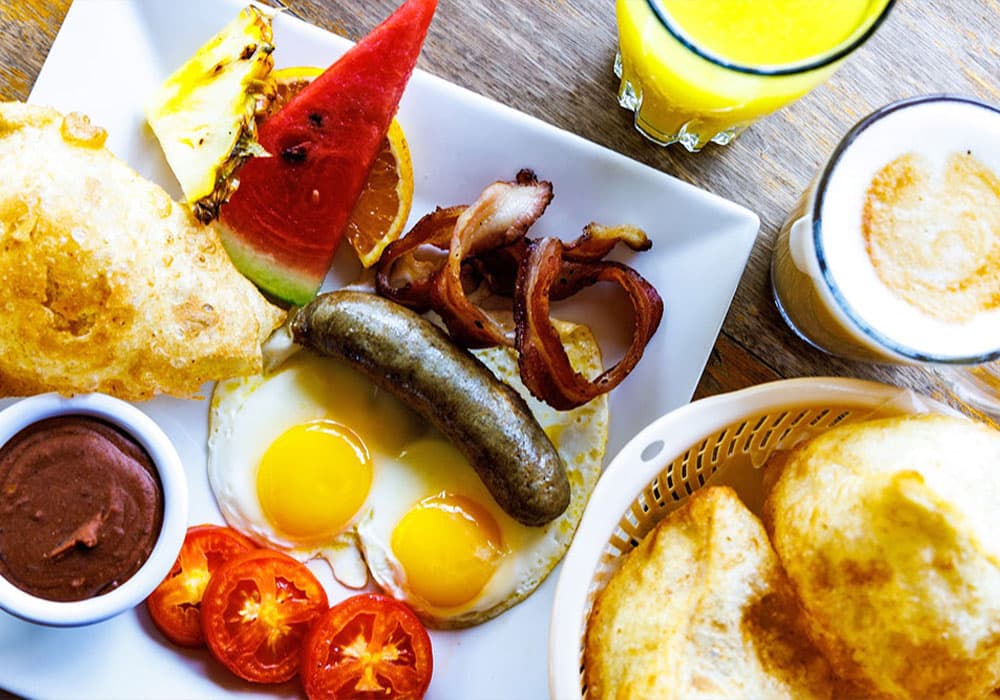 A colorful breakfast plate featuring eggs, sausage, bacon, tomatoes, fresh fruit, and drinks.
