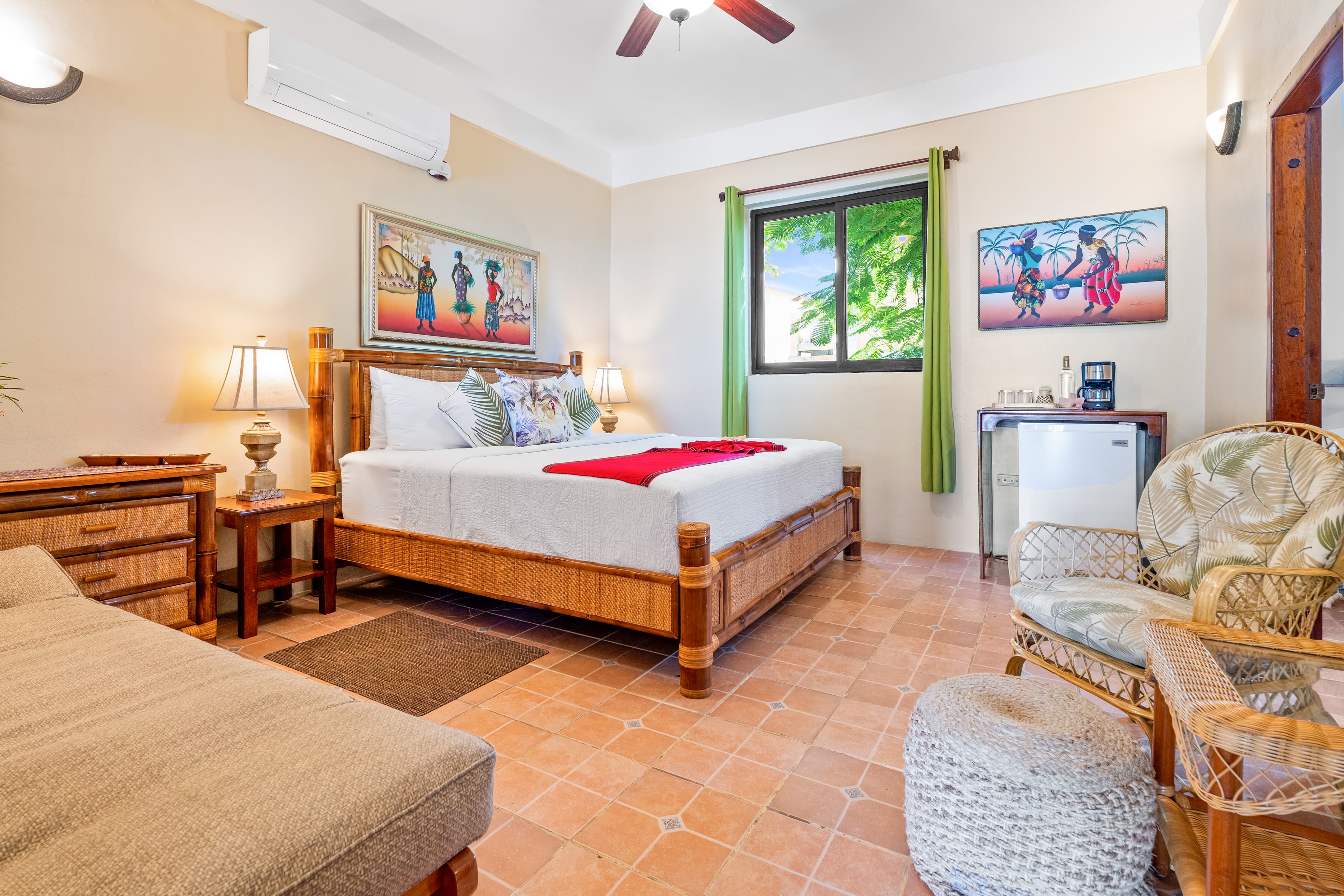 A spacious bedroom with terracotta tile flooring and light beige walls, featuring a king-size bamboo bed with white linens and a vibrant red runner. The room is furnished with a tan chaise lounge in the foreground, a bamboo dresser, and a seating area with a wicker chair and a woven ottoman. Large windows with green curtains provide a view of lush trees, and several pieces of colorful tropical artwork are displayed on the walls.