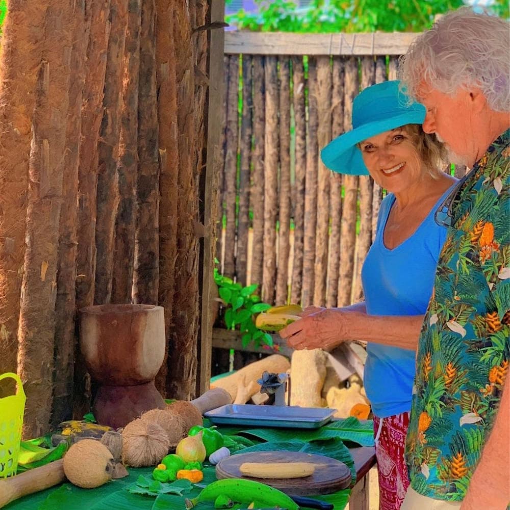 Two individuals, a woman in a blue shirt and a hat, and a man in a patterned shirt, are preparing food at a table filled with various fruits and vegetables in a lush outdoor setting.