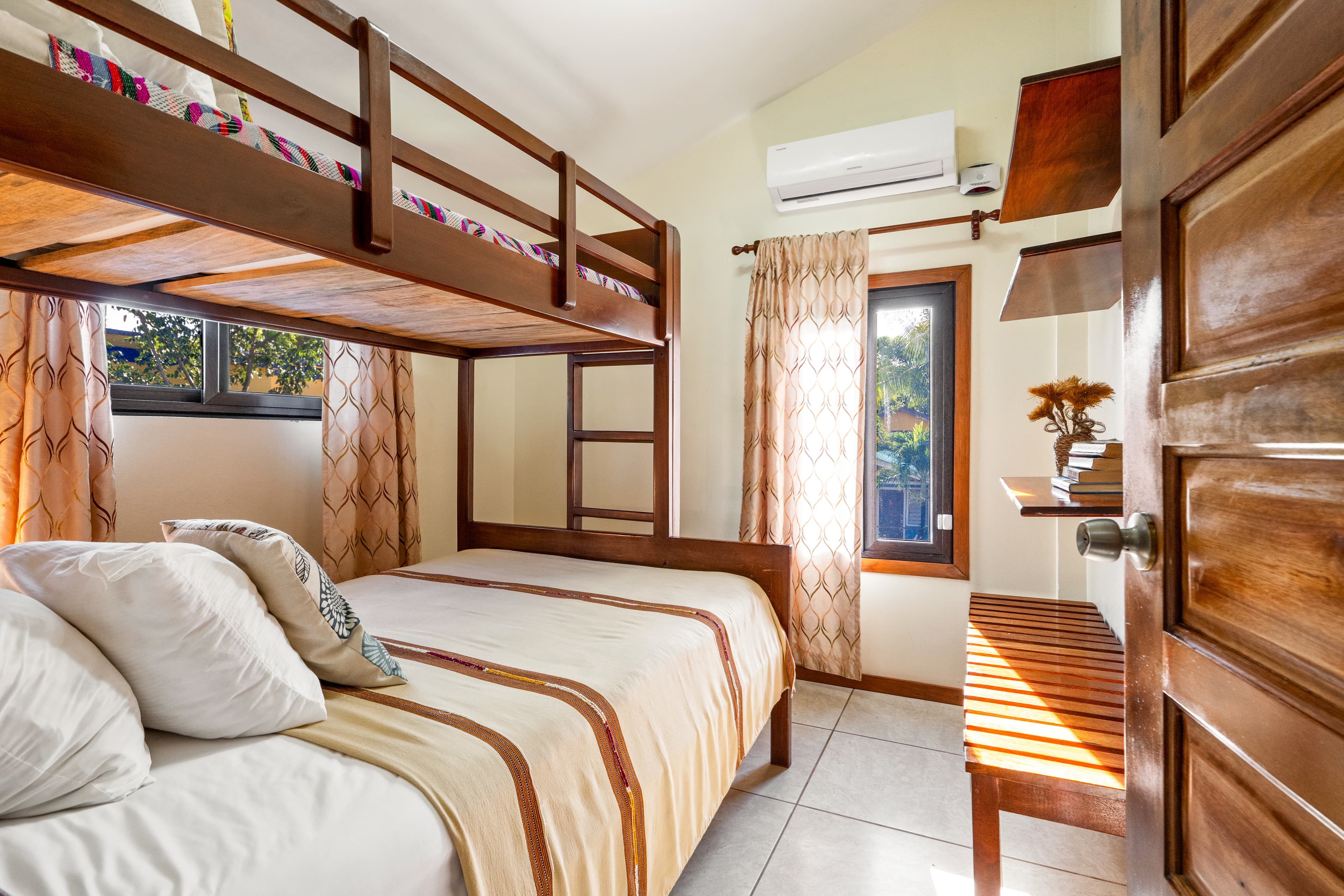A bedroom featuring a wooden bunk bed with white linens and a striped runner, positioned next to two windows framed by patterned beige curtains. The space includes a wooden bench, wall-mounted shelves, and a view into the room through an open wooden door.