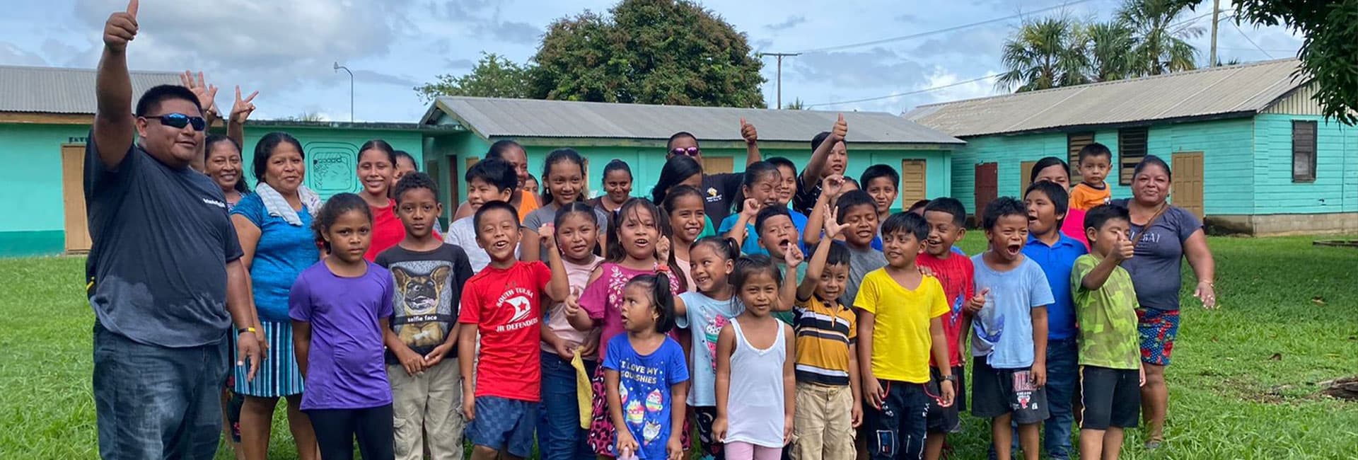 A large group of children and adults stands outside, smiling and giving thumbs up in front of green buildings.