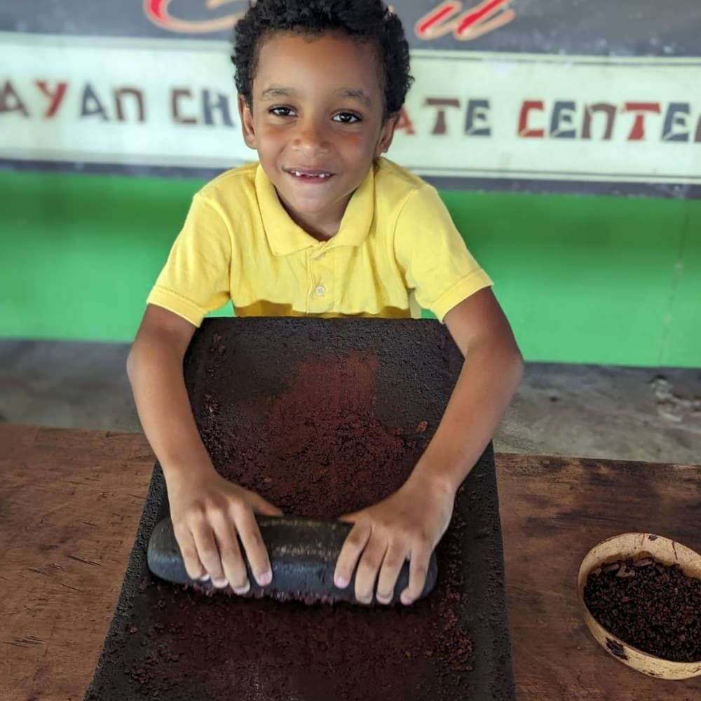 A smiling boy in a yellow shirt uses a rolling stone on cocoa beans at a chocolate center.