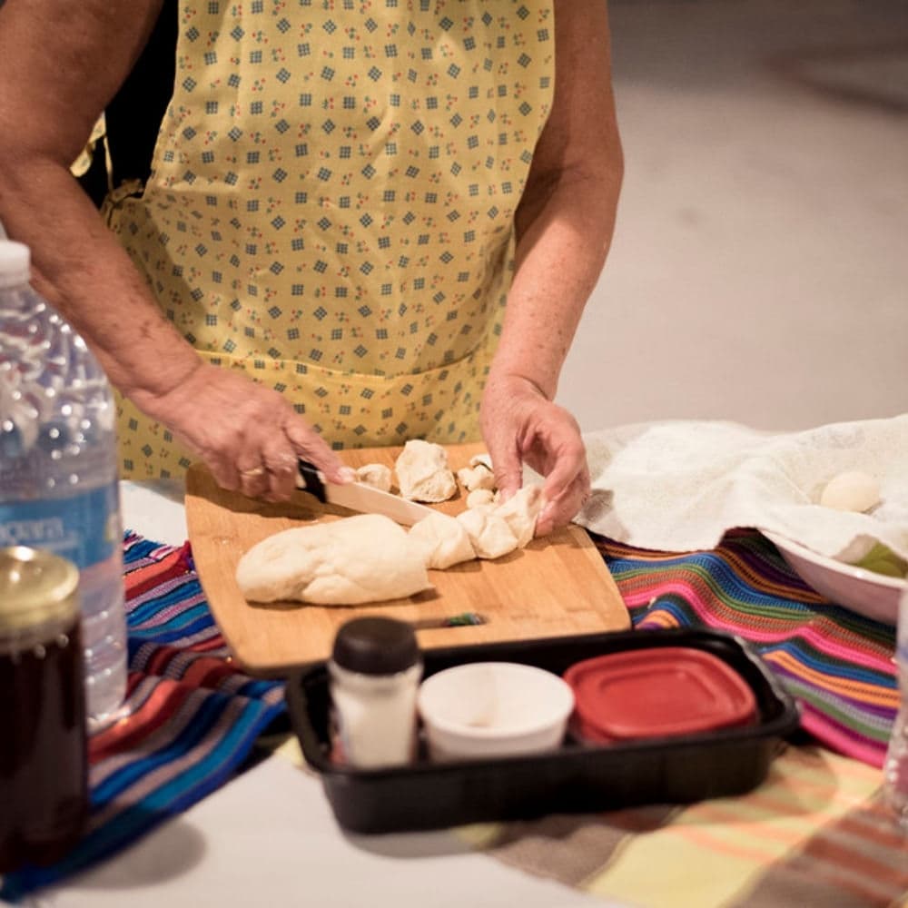 An individual in a yellow apron chops dough on a wooden cutting board, surrounded by food items and a colorful tablecloth.