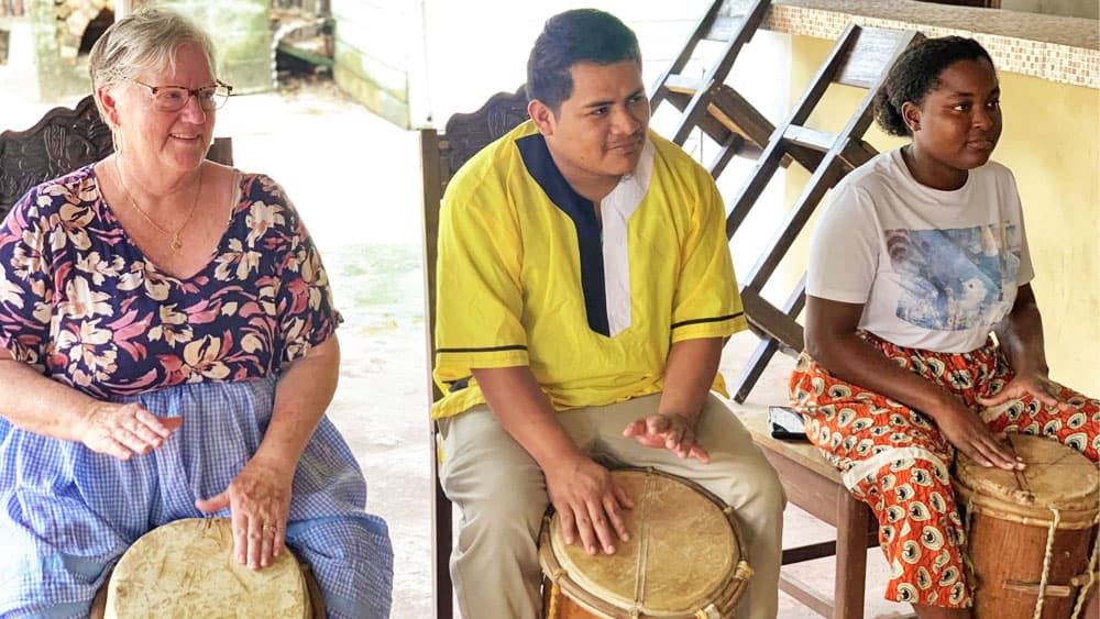 Three individuals play drums together, smiling and engaging in a musical activity in a relaxed setting.