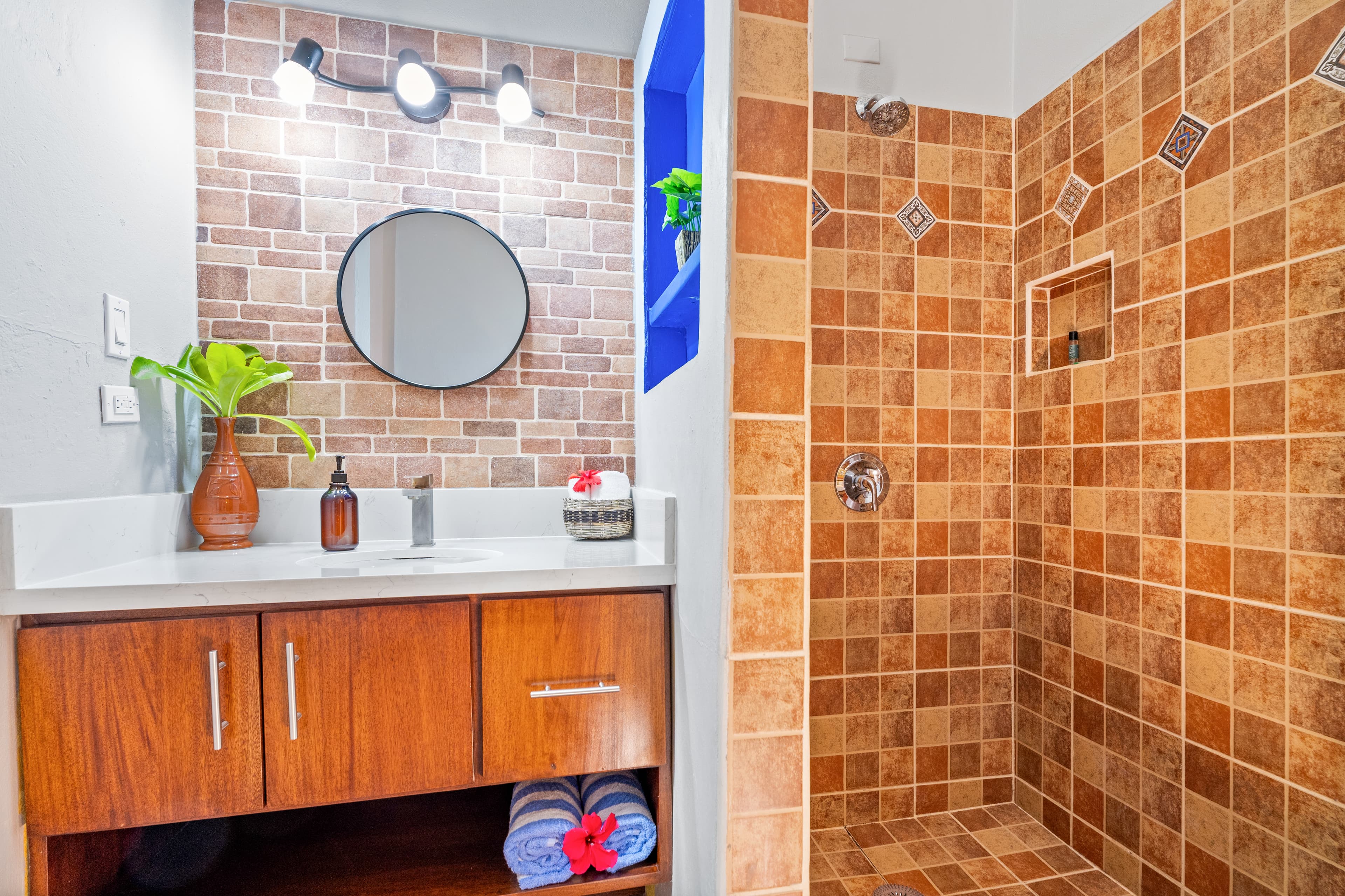 A modern bathroom with warm brown tiles featuring a wood-grain vanity with a white countertop and a circular mirror. To the right is a walk-in shower with matching textured brown wall tiles and a small built-in shelf. The vanity is decorated with an orange vase containing a green plant, a small woven basket, and blue striped towels tucked into the open shelf below.
