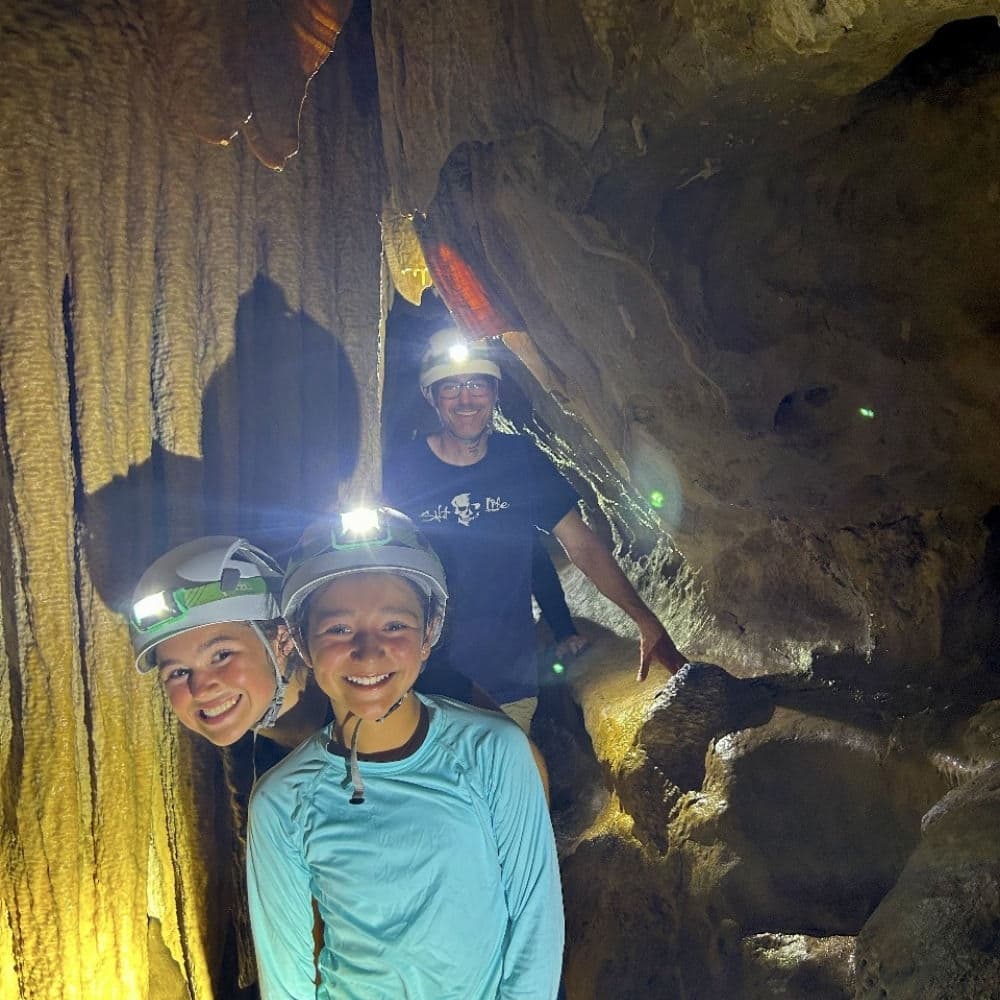 Three people, wearing helmets with headlamps, smile in a cave surrounded by rock formations.