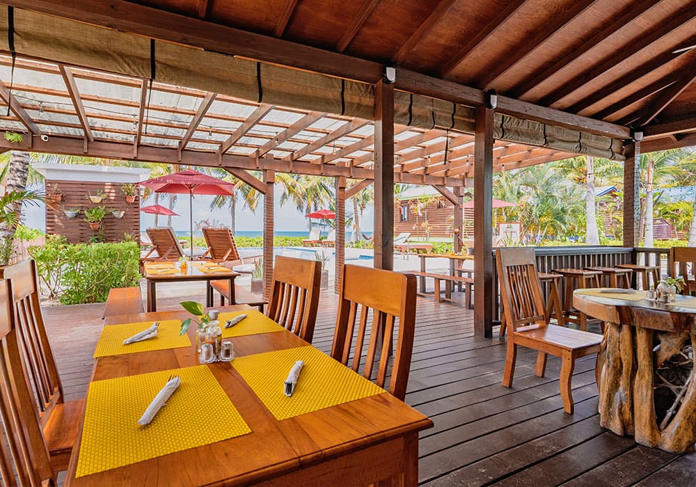Interior view of a tropical restaurant with wooden tables and a patio overlooking a pool and palm trees.