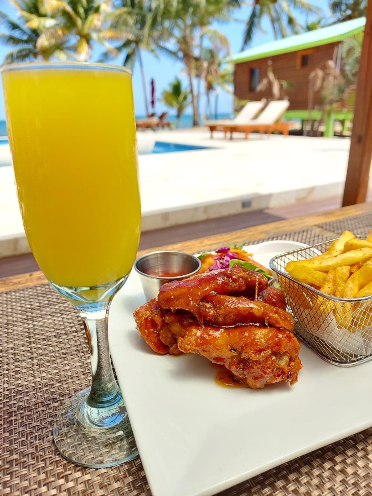A plate of spicy chicken wings and fries accompanied by a glass of yellow beverage, set against a tropical backdrop.