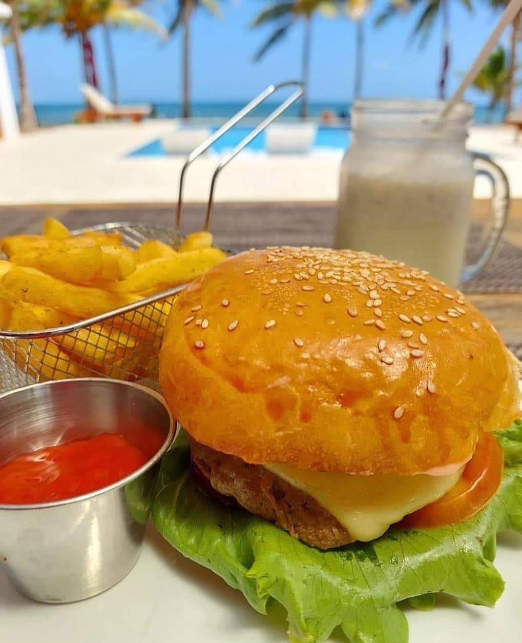 A sesame-seed hamburger with lettuce, cheese, and a side of fries and ketchup, set against a beachside backdrop.