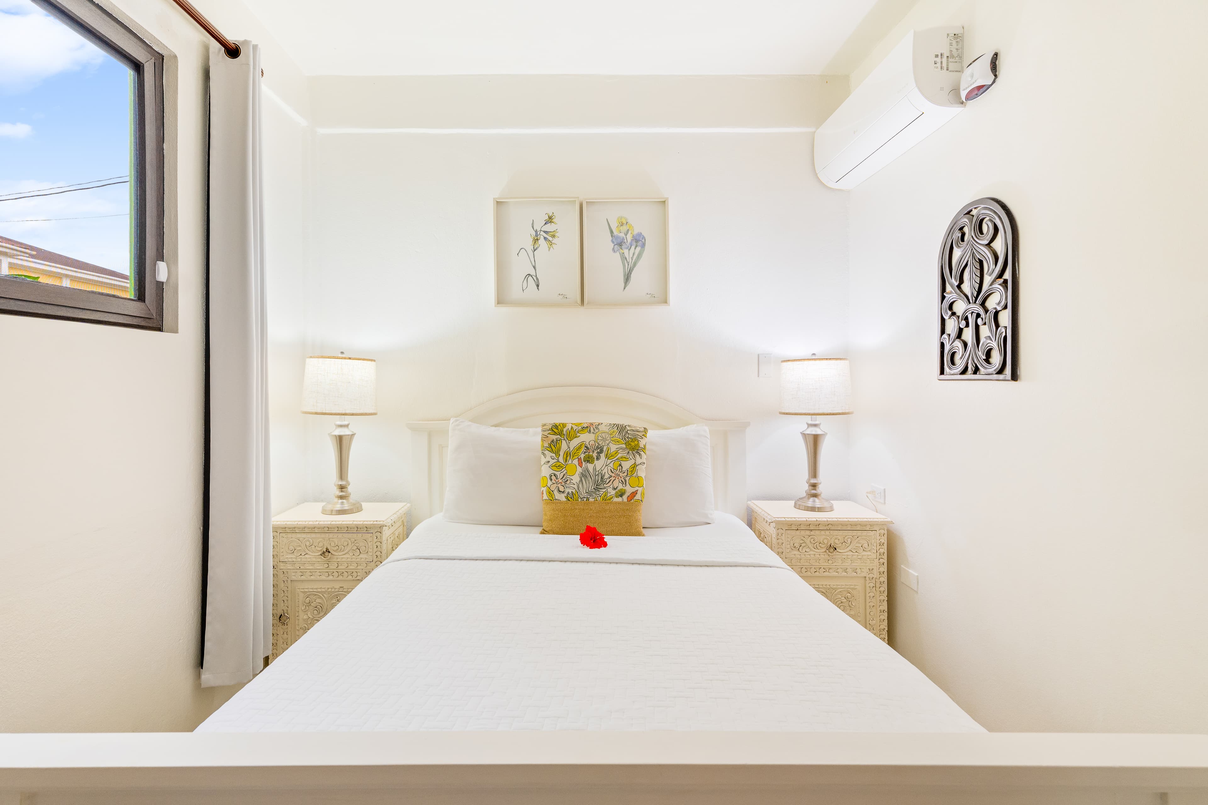 A brightly lit, minimalist bedroom featuring a white-sheeted queen bed with a yellow floral accent pillow and a single red flower. The room includes matching white nightstands with lamps on either side of the bed, two small botanical art prints on the wall, and a large window to the left.