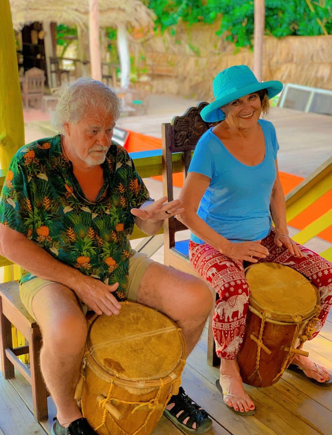 Two people play hand drums while sitting on chairs in a tropical setting.