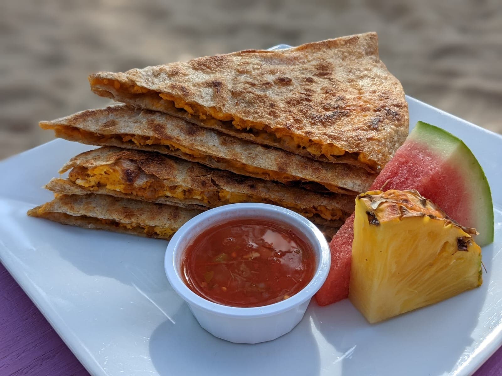 A plate with stacked cheese quesadillas, a side of salsa, and fresh fruit including watermelon and pineapple.