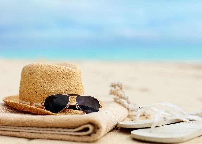 A straw hat, sunglasses, and white flip-flops resting on a sandy beach towel with a blurred seascape in the background.